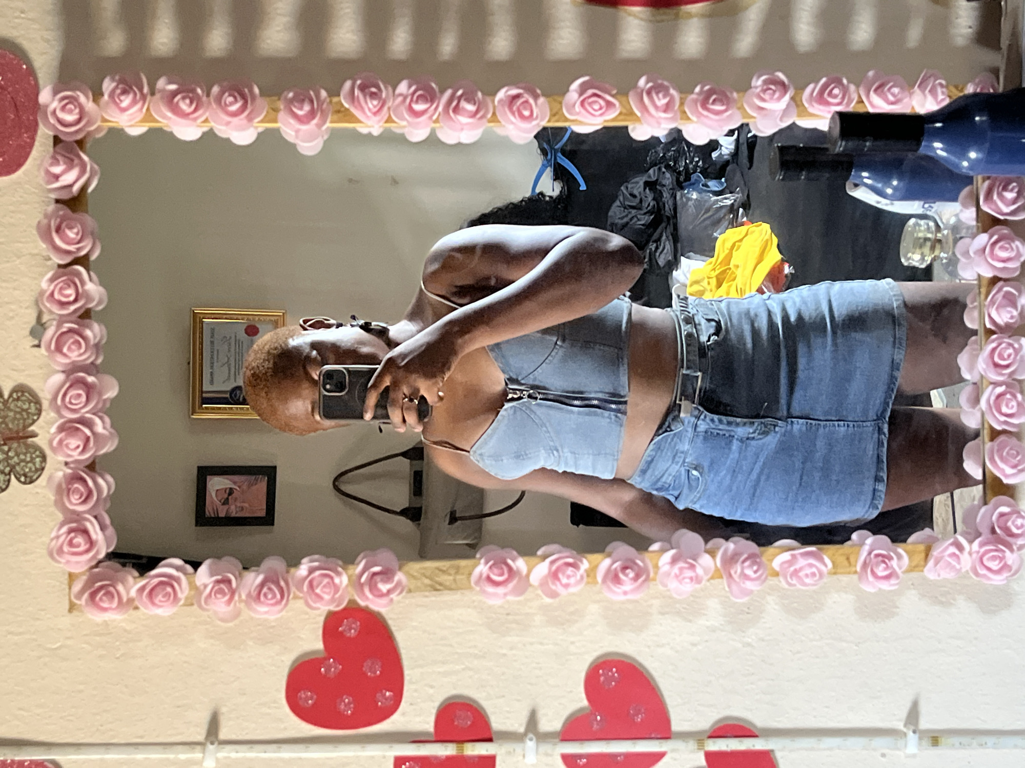 An individual is captured in a mirror selfie, with the image rotated 90 degrees clockwise. The subject, a person with short, reddish-brown hair, is holding a smartphone to take the photo, partially obscuring their face. They are wearing a light denim or chambray-like bustier top with a zipper front and a matching denim mini-skirt.
The selfie is taken in a rectangular mirror, framed by decorative pink rose-shaped embellishments. Reflected in the mirror's background are several items on a light-colored wall, including a gold-framed certificate or document, and a smaller black-framed picture below it. Other reflected objects include a black plastic bag, a yellow garment, a blue bottle, a clear glass bottle, and various cables, indicating a potentially cluttered indoor setting.
Outside the mirror's frame, on the wall, there are several red heart-shaped decorations, some with glitter, along with a light-colored butterfly decoration. The overall aesthetic suggests a personal, decorated space, possibly a bedroom or dressing area. No specific time of day or discernible emotion is evident. The general lighting suggests it is daytime. The location context provided is Jalingo, Nigeria. Visible text on the reflected certificate is largely illegible.