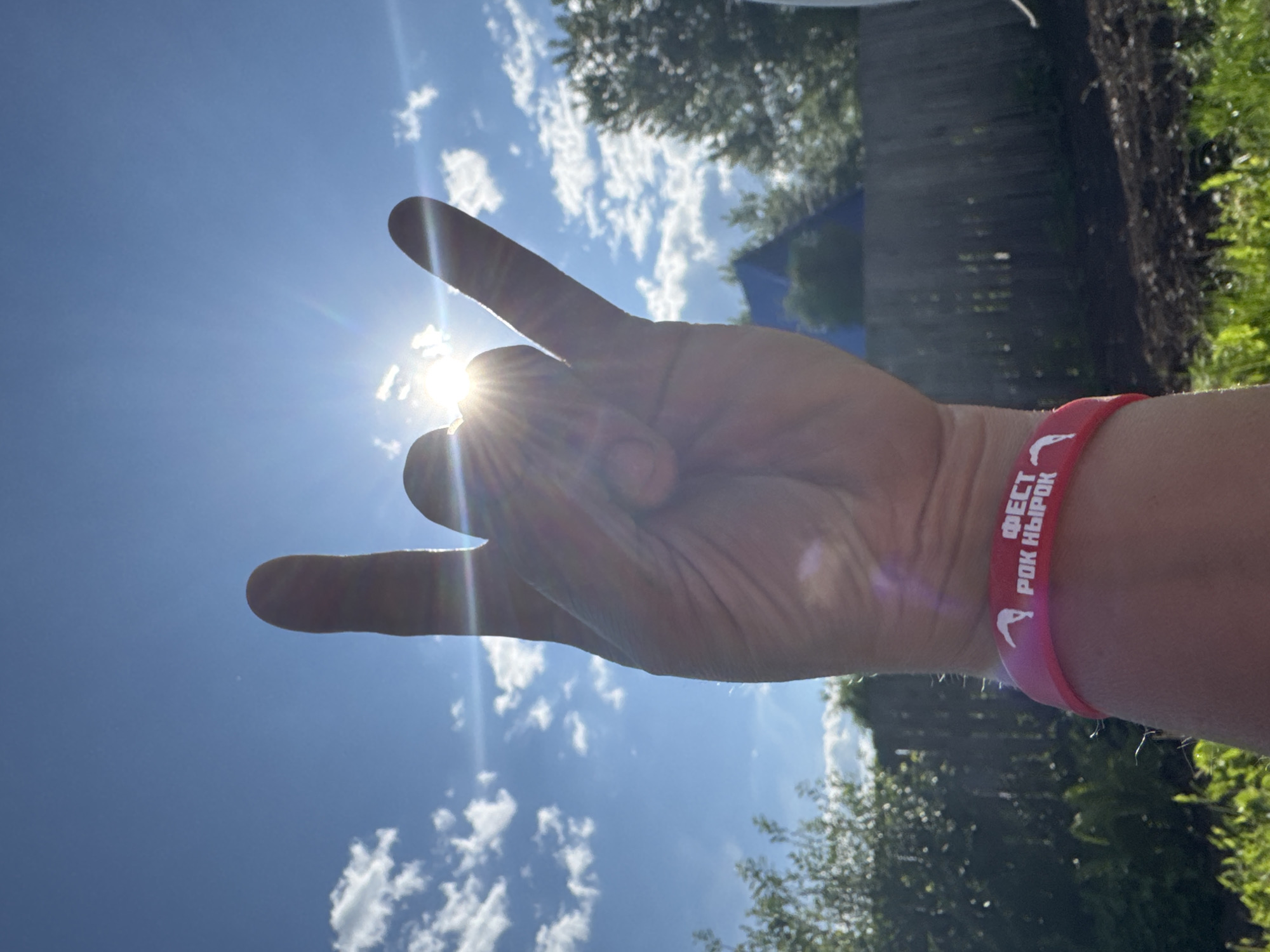 The image displays a human hand making the "rock on" gesture (corna), silhouetted against a bright blue sky. The hand is positioned centrally in the foreground, with the index finger and pinky extended upwards, and the middle and ring fingers curled and held by the thumb. Direct sunlight emanates from behind the hand, specifically from the upper right quadrant, creating a strong sunburst effect and lens flares that partially obscure the hand's details.
A pink silicone wristband is visible on the subject's left wrist. The wristband features white Cyrillic text that reads "ФЕСТ РОК НЫРОК," which translates to "Fest Rock Dive" or "Rock Plunge Fest." Adjacent to the text is a white graphic depicting a stylized figure in what appears to be a diving or acrobatic pose.
The background consists of a clear, bright blue sky with scattered white cumulus clouds, predominantly in the upper left section. The lower third of the image shows an outdoor environment. On the left, dense green foliage, consistent with bushes and trees, is visible. In the mid-ground, a wooden fence, possibly a picket fence, runs horizontally. Behind the fence, a dark blue, pointed structure, resembling a tent or a roof, is partially visible among more green trees. The ground in the immediate foreground features green grass mixed with patches of darker, tilled earth. The scene is brightly illuminated, indicating a sunny day. This outdoor scene is located in the city of Chaikovsky, Russia, as indicated by the Cyrillic text on the wristband.