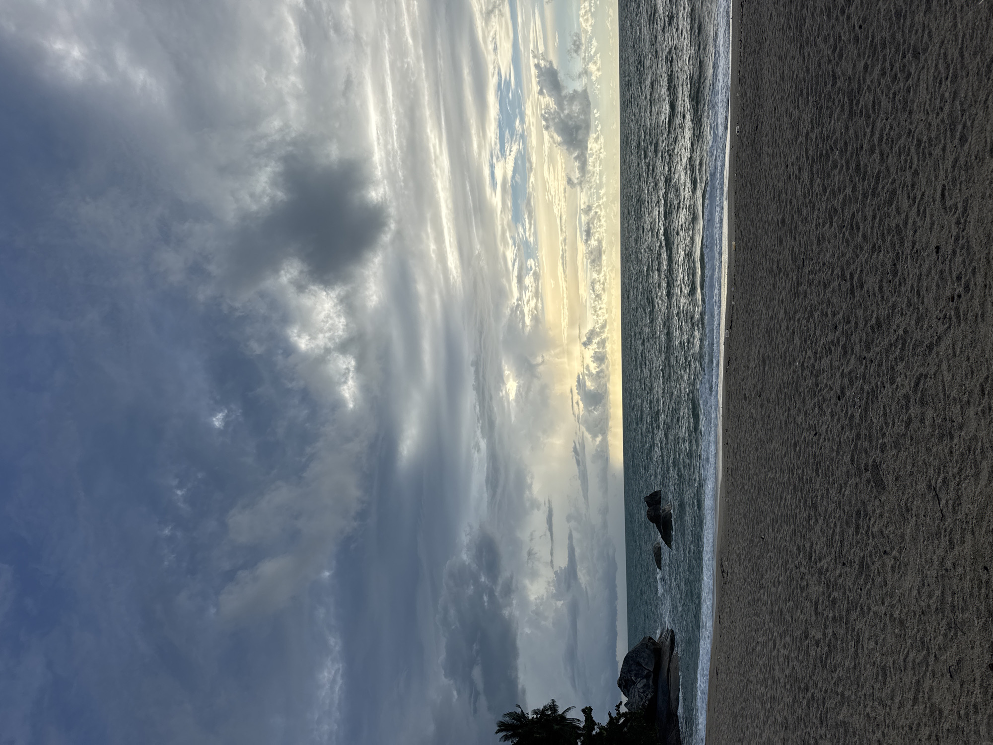 The image depicts a beach scene in Baan Thong Nang, Thailand. The sky is filled with striated clouds, ranging from dark gray to white, with some patches revealing the blue sky. A section of the sky near the horizon has a yellow cast, likely due to the sun. The ocean is to the right of the sky, the water appears to be a dark, rippled expanse. Adjacent to the ocean is a wide expanse of dark sand. In the lower portion of the image, near the shoreline, there are some rocky formations and some foliage, possibly palm trees.