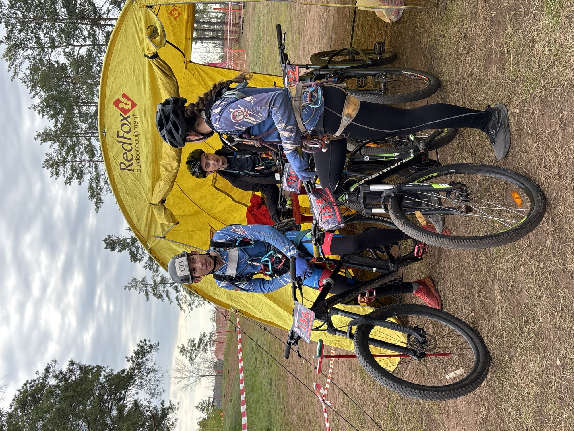 The image shows three people standing near a yellow "RedFox outdoor equipment" tent in an outdoor setting. Two are wearing helmets and blue cycling attire with harnesses, leaning on mountain bikes. The man on the left wears a helmet with the number "193". The other person, possibly a female, has braided hair and a black helmet. A third person in a black jacket is standing behind them. All are presumably preparing for or have just finished a cycling or sporting event. The location appears to be a grassy area with trees and a striped barrier visible in the background. Based on additional information, the city is Orenburg, Russia.