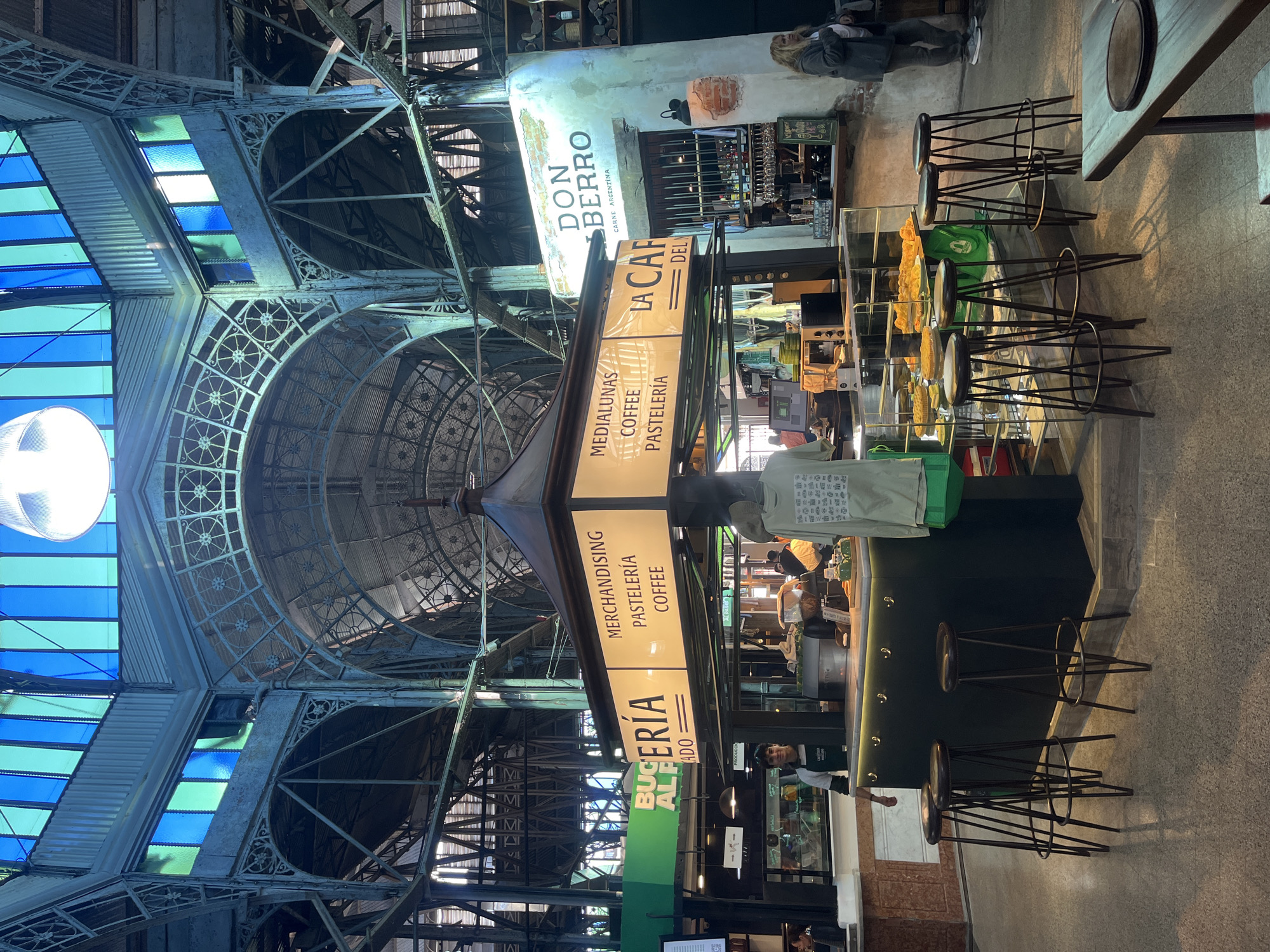 The media file presents an interior view of a vibrant market or food hall, identified by architectural cues and visible text as being in Buenos Aires, Argentina. The scene is dominated by a large, ornate metal structure with an arched, almost dome-like ceiling and intricate filigree patterns. Sections of blue and light green translucent panels in the roof and upper walls allow natural light to filter in, suggesting it is daytime.
In the center, a multi-sided kiosk-style counter serves as a focal point, specializing in pastries and coffee. Vertical signs prominently display "MEDIALUNAS," "COFFEE," and "PASTELERÍA," alongside "MERCHANDISING," "PASTELERÍA," and "COFFEE," with a partially visible sign that reads "LA CAFETERÍA DEL...". Below these signs, glass display cases showcase a variety of baked goods, such as tarts or quiches. Several dark-toned bar stools with metal frames are arranged around the counter, indicating seating for patrons. Staff members are visible behind the counter, engaged in serving customers.
Further into the background, another establishment features a rustic, whitewashed wall with a sign reading "DON BERRO CARNE ARGENTINA," suggesting a meat vendor. Other people, appearing to be both customers and additional staff, are present within the bustling environment. The flooring is a light, neutral color. The overall atmosphere is lively and casual, characteristic of a popular food destination.