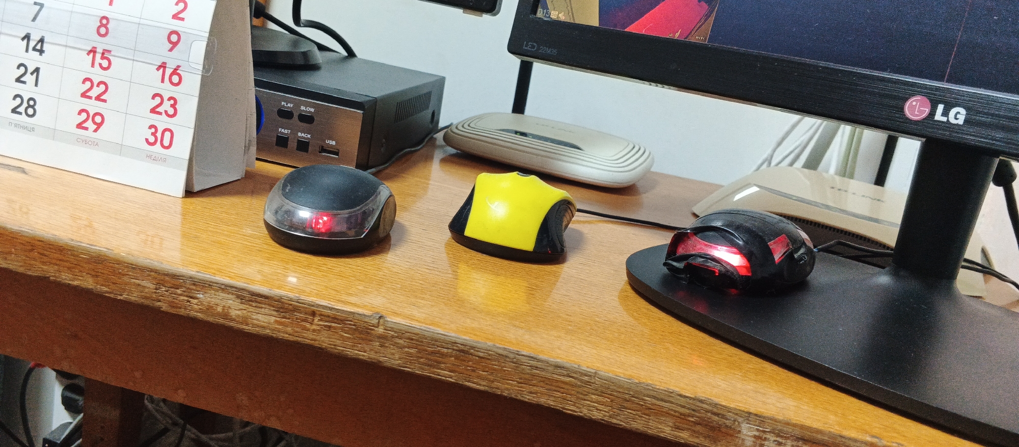 Three computer mice are positioned on a wooden desk. The mouse on the left is black with a clear plastic top that has a red LED light glowing. The middle mouse is yellow and black. The mouse on the right is black with red LED lights. To the left of the mice is a calendar, displaying the numbers 7, 8, 9, 14, 15, 16, 21, 22, 23, 28, 29, and 30. Behind the calendar is a black electronic device with buttons labeled "PLAY", "SLOW", "FAST", and "BACK", and a USB port. To the right of the electronic device is a white, curved electronic device. Further to the right, a black computer monitor stands on a black stand. The monitor has an LG logo on its front bezel. The overall environment suggests a home or office workspace. The image is captured from a close-up perspective, focusing on the desk and its contents. There is no visible indication of the city of Smila, Ukraine, within the image itself.