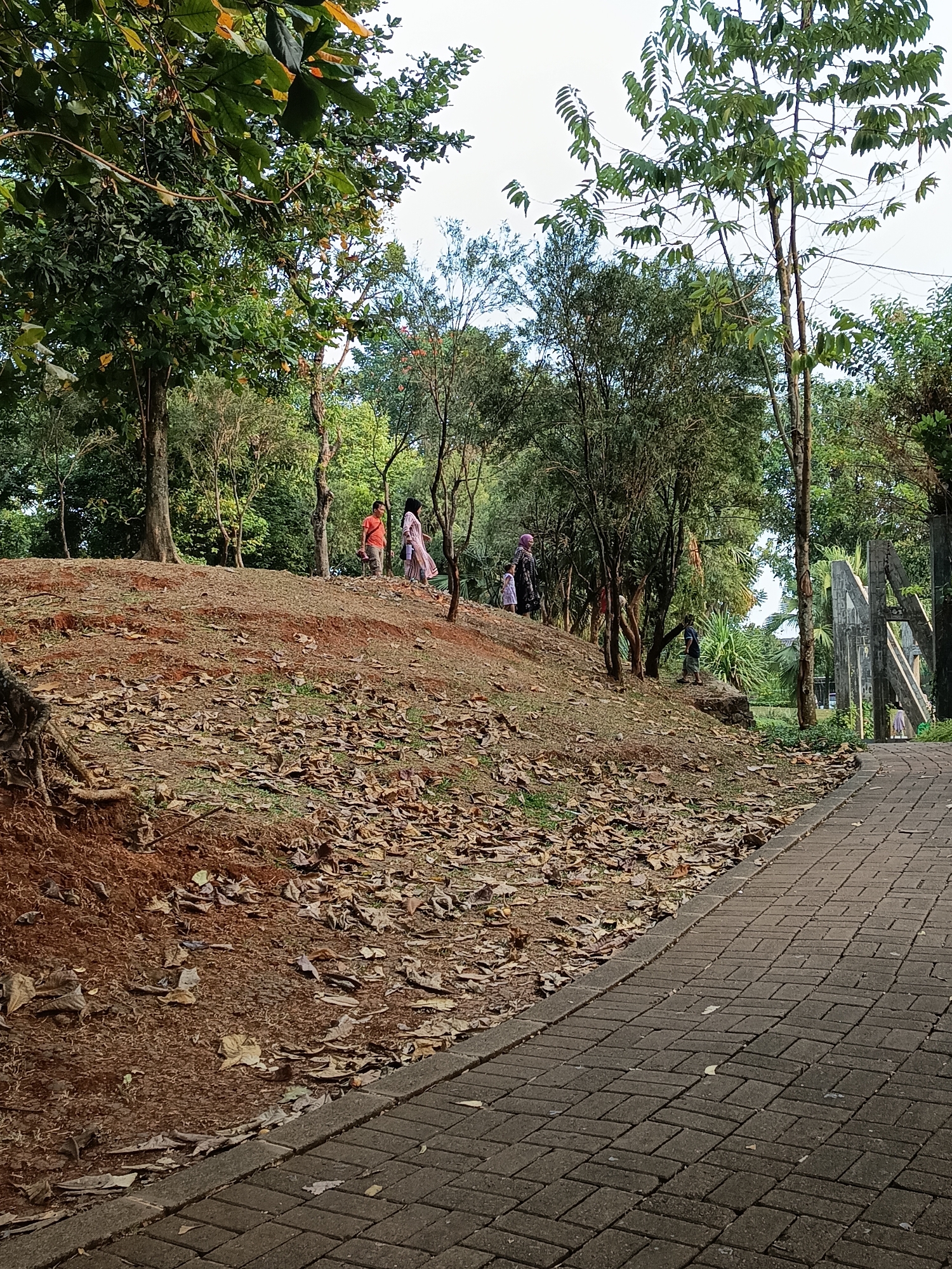 The image depicts a park scene in East Jakarta, Indonesia, on a bright day. In the foreground, a paved walkway made of dark gray rectangular bricks runs diagonally from the bottom right corner towards the center of the image. The walkway is bordered by a strip of brown earth covered in dry, fallen leaves. To the left of the walkway, a sloping mound of reddish-brown earth is also covered with a dense layer of fallen leaves. The mound is covered with sparse green grass.
Further into the background, several trees with green foliage are scattered across the landscape. On the mound, at various distances, are several individuals. One person in an orange shirt is visible walking uphill, while another person in a light-colored, patterned dress is walking uphill beside them. Two other individuals, one in a dark shirt and the other in a lighter colored dress, are also on the mound. In the far background, a large, abstract concrete structure with a geometric design stands to the right of the trees. The sky above is bright and appears to be clear.