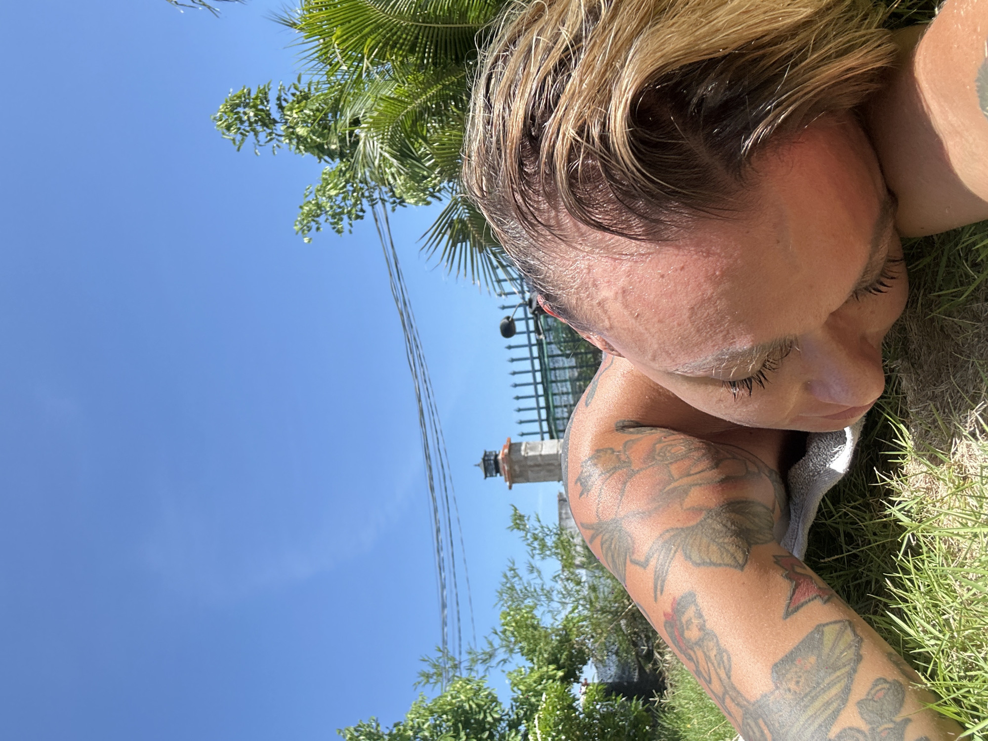 A close-up, ground-level shot depicts a person lying on grass outdoors. The individual has fair skin, damp blond hair, and extensive tattoos covering their right arm. The camera angle is from a low perspective, capturing the person's face in profile, with their eyes closed.
In the background, a clear blue sky is visible, along with the tops of trees and a metal fence. Wires and a concrete lighthouse are partially visible, suggesting the location is near a coastal area. Based on the prompt, the location is Mũi Né, Vietnam, near the Ke Ga Lighthouse.