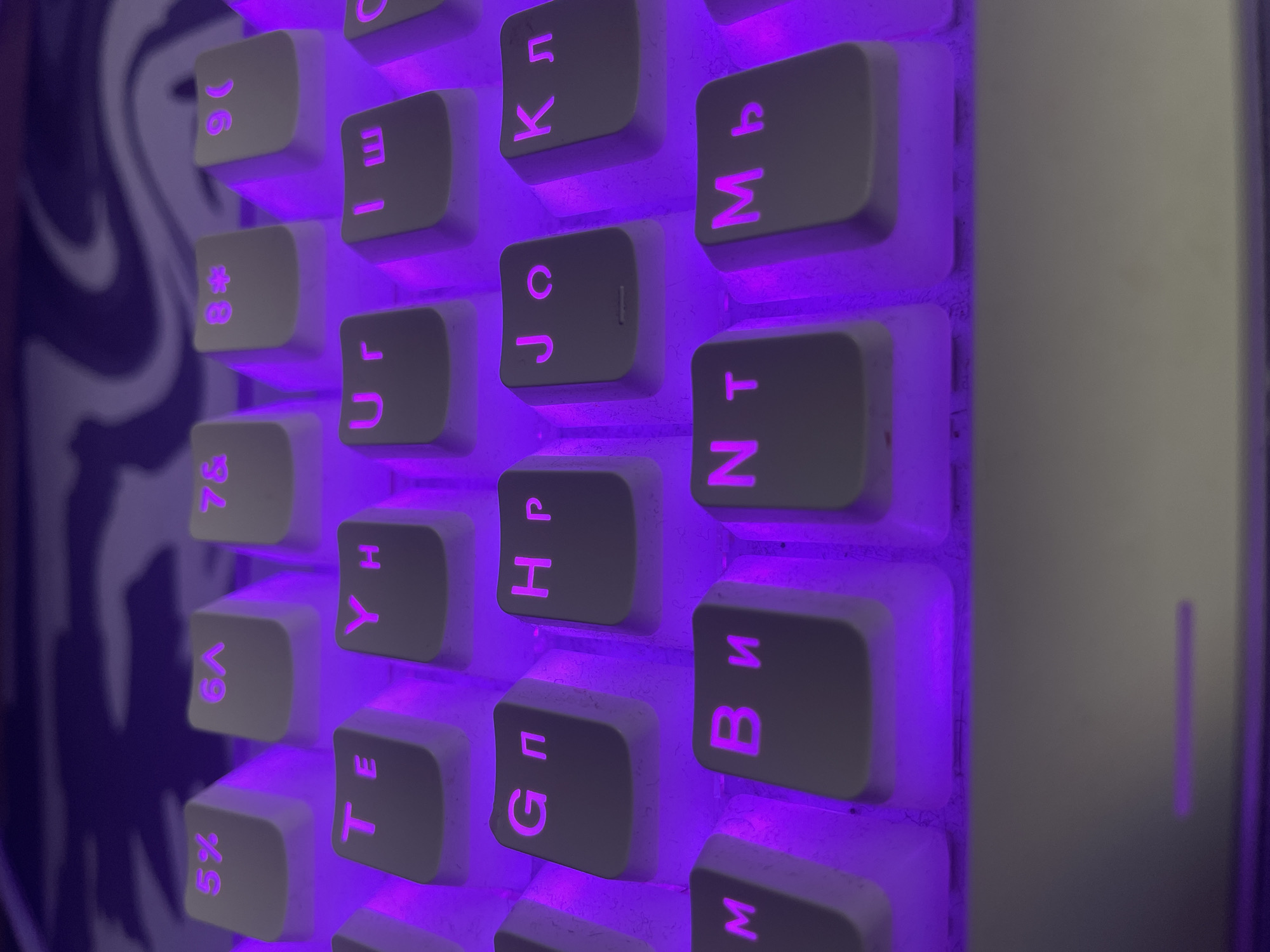 The image is a close-up, top-down view of a white mechanical keyboard with purple backlighting. The keyboard features a mix of English and Cyrillic characters on its keys. Visible keys include "5%", "6^", "7&", "8#", "9(", "T", "E", "Y", "H", "U", "r", "I", "ш", "G", "п", "H", "p", "J", "o", "K", "л", "M", "B", "и", "N", "T", "M", "ь". The background appears to be a patterned surface, possibly a desk mat, with swirls of white and grey. The lighting is dim, with the purple glow of the keyboard illuminating the surrounding area. There are no people present in the image, and no specific location within Sinyavino, Russia, can be identified. The scene is static and focuses solely on the keyboard.