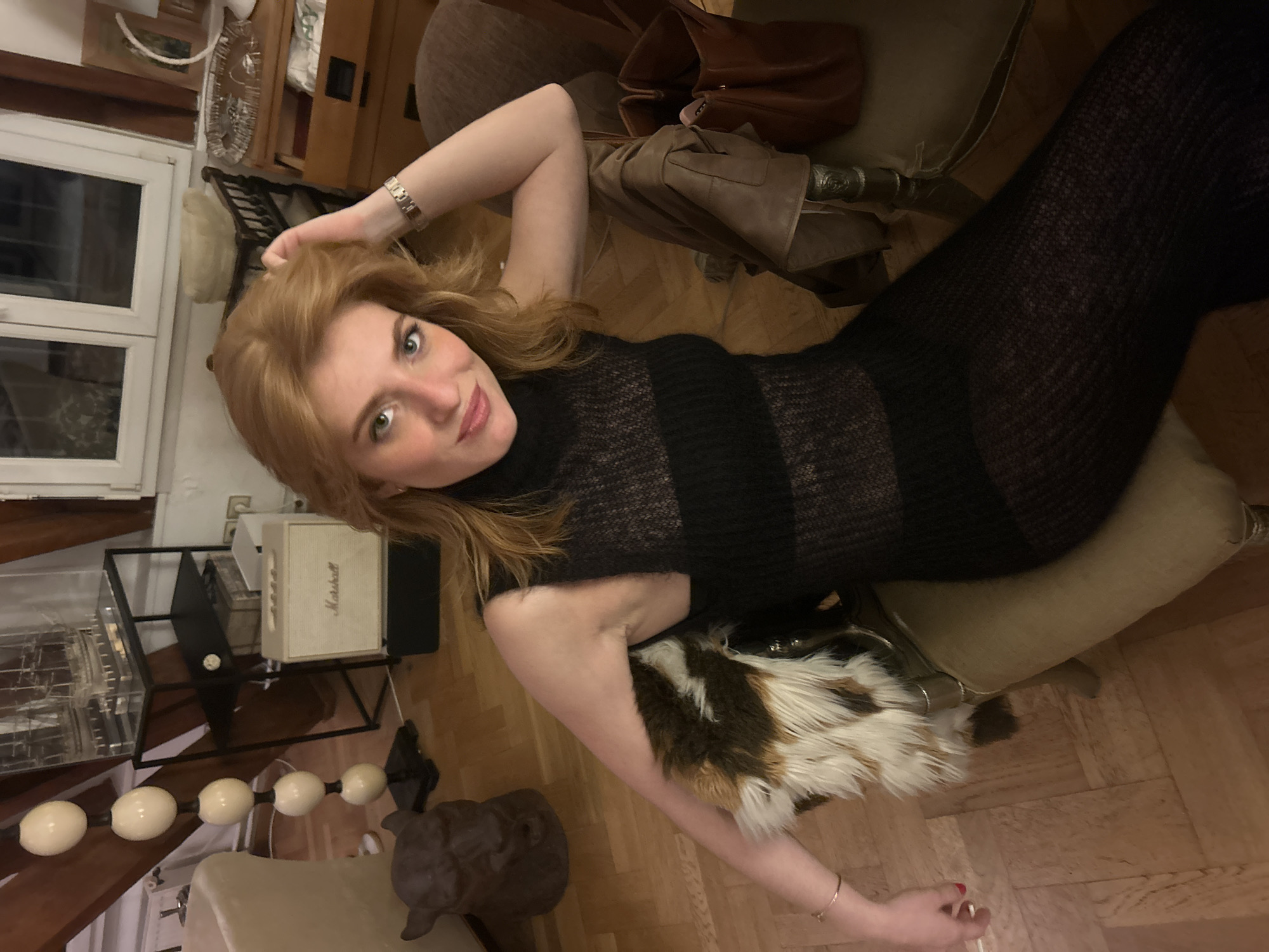 A woman lies on a light-green chaise lounge. She is wearing a black semi-sheer turtleneck sweater dress. She has red hair. Her left arm is raised above her head. Her right arm is lying on a fur blanket on the floor. There are several objects in the room, including a Marshall speaker, a glass display case, and a wooden cabinet. The room has wooden floors. The location is a private residence in an unknown city.