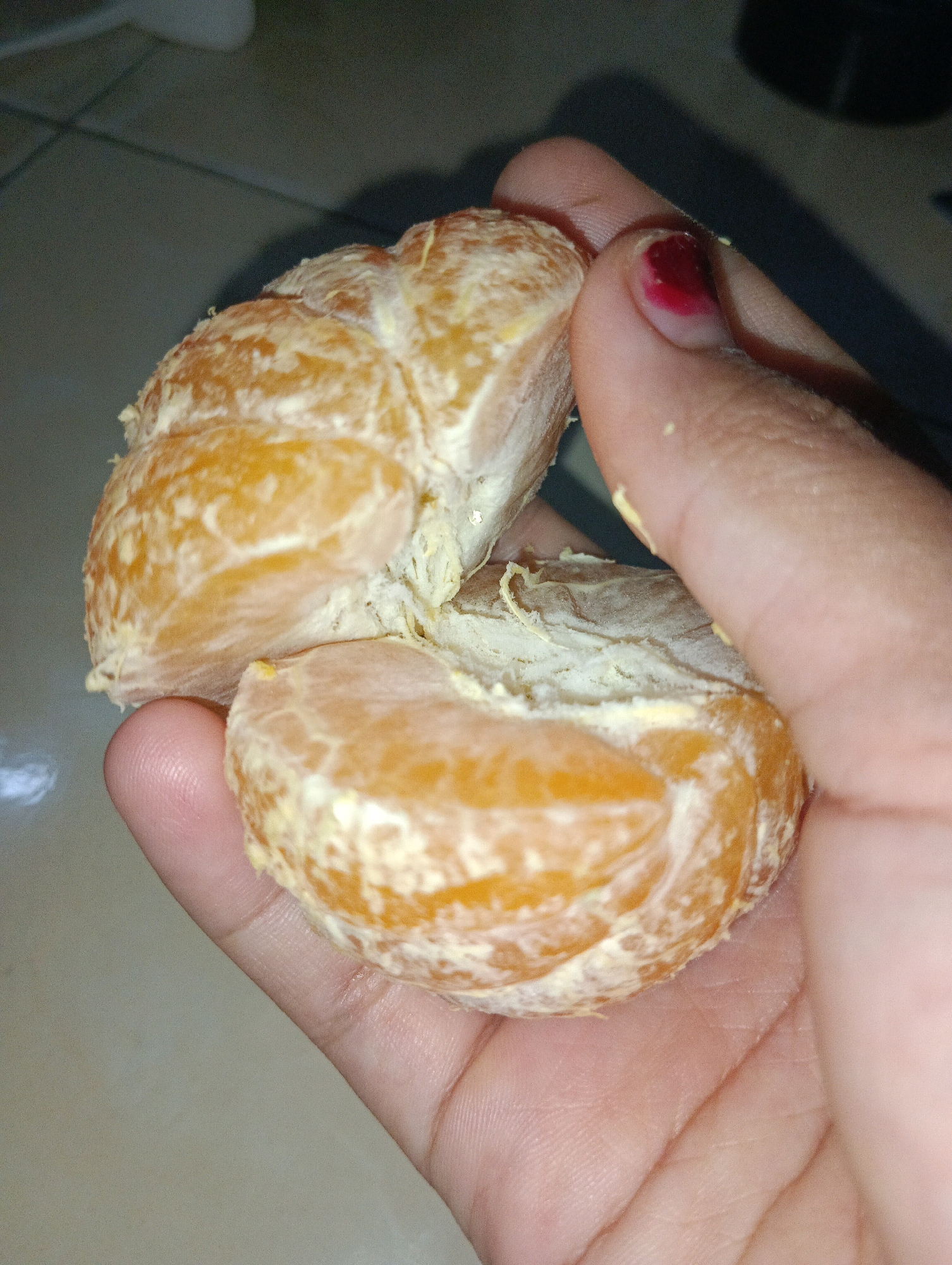 A person's hand, with a dark red nail polish on the thumbnail, is holding a partially peeled tangerine. The tangerine is bright orange and divided into segments, with some of the white pith still attached. The background appears to be a tiled surface, possibly a floor or countertop, in a dimly lit room. The image is a close-up, focusing on the tangerine and the hand. There are no discernible landmarks to indicate a specific location in Pematang Jaya, Indonesia.