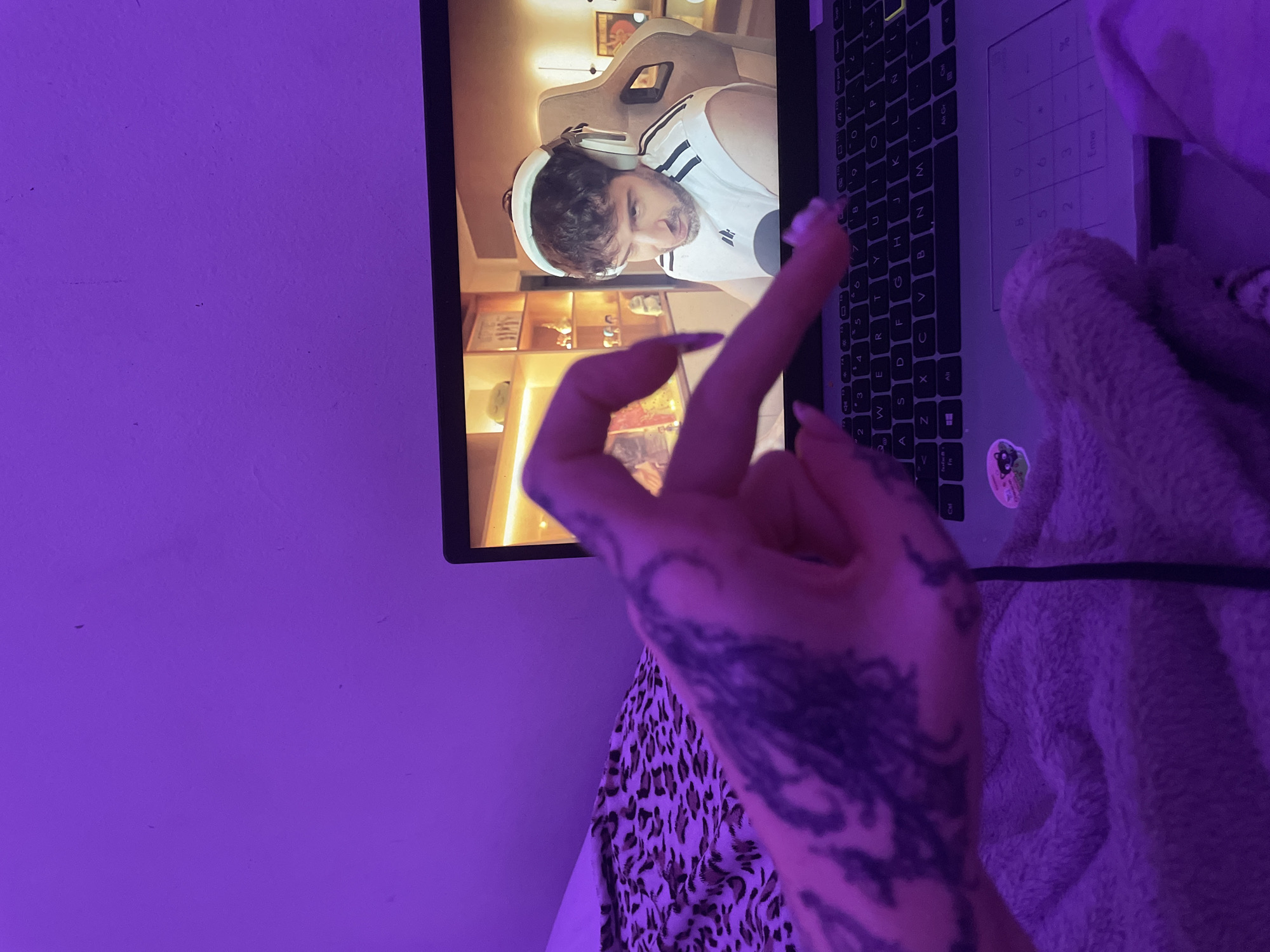A laptop screen shows a man wearing headphones and an Adidas tank top in front of a tan gaming chair, seemingly indoors. A hand with a tattoo on the arm is extending the middle finger in front of the laptop. The hand has long, pointed fingernails. Part of the laptop keyboard is visible as well as part of a fluffy blanket or sweater. The image is lit with a purple light. The location in Buenos Aires, Argentina cannot be determined from the image.