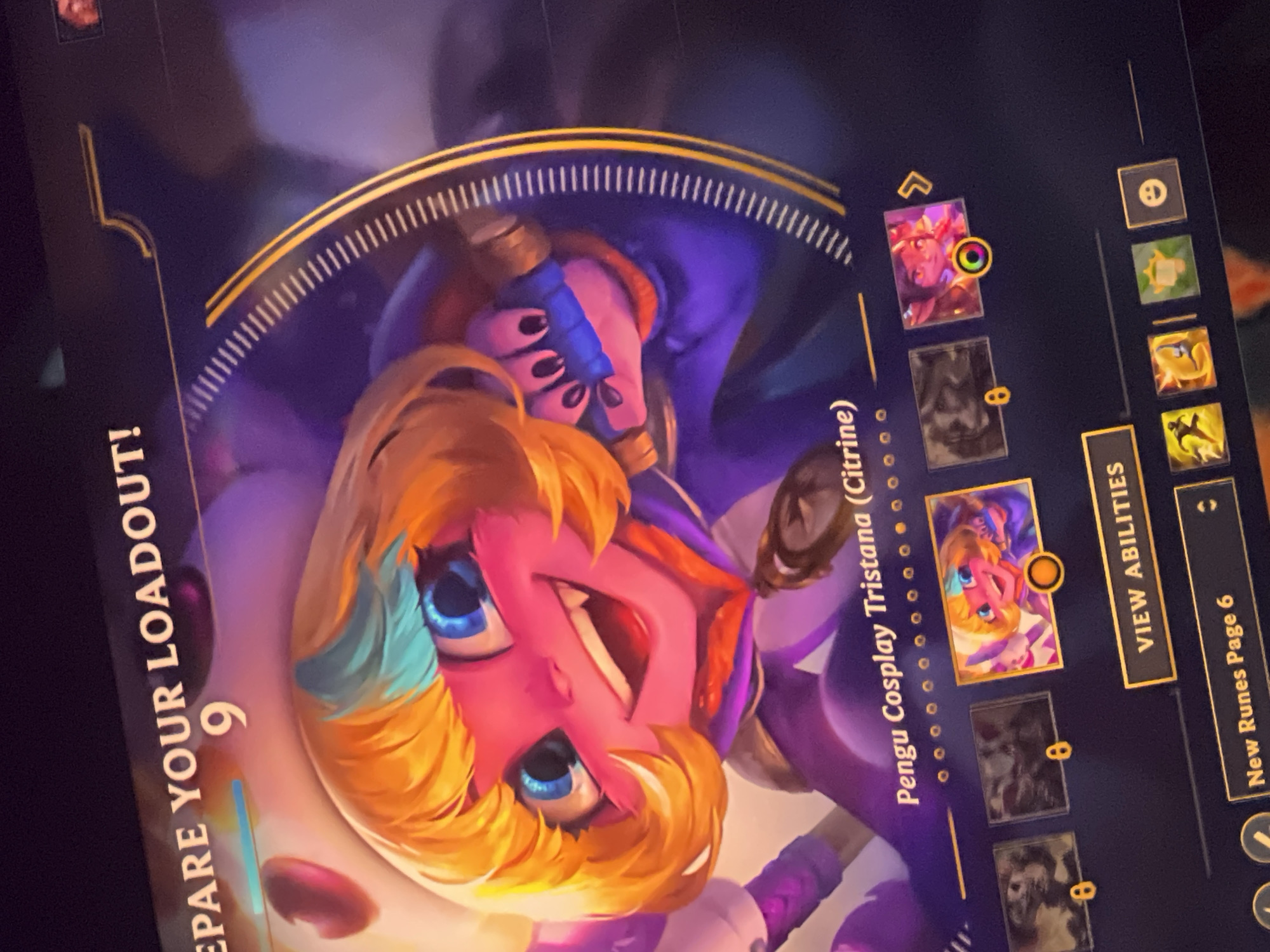 This is a close-up, angled, eye-level photograph of a video game interface displayed on a screen. The main focus is a character portrait of a cartoonish female character with bright pink skin, large blue eyes, and blonde hair with a turquoise streak. She has an excited expression with a wide grin revealing pointed teeth. The character is depicted in a purple outfit with gold accents. Above the character portrait, text reads "EPrepare YOUR LOADOUT!" with the number "9" prominently displayed. Below the character portrait is the text "Pengu Cosplay Tristana (Citrine)". A row of smaller icons is visible at the bottom of the screen, along with buttons labeled "VIEW ABILITIES" and "New Runes Page 6". The background is dark and blurred, suggesting it is part of the game's visual environment. The lighting appears to be from the screen itself, creating reflections.
