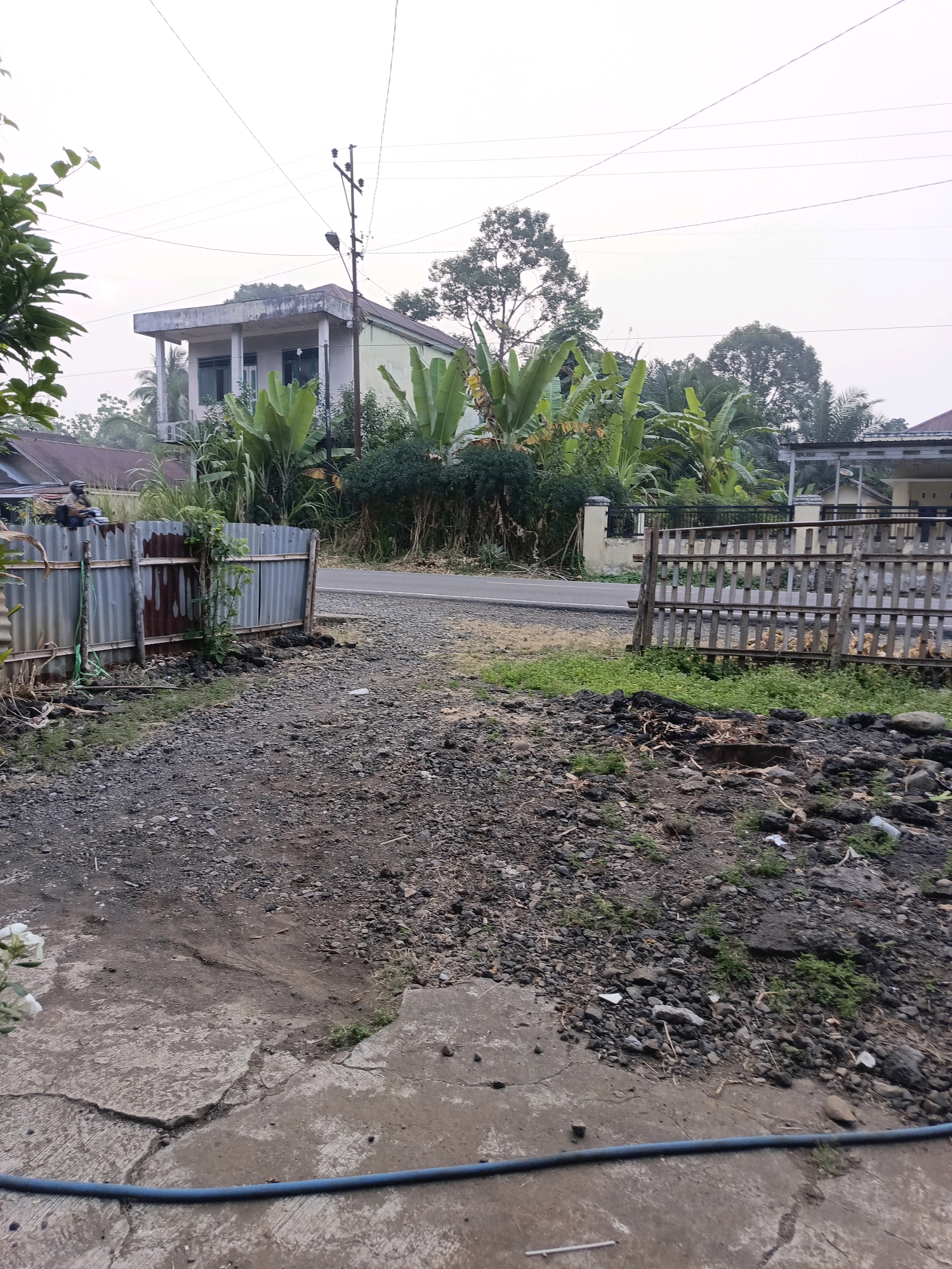 The image depicts a street scene in Ulak Tanding, Indonesia, characterized by a paved road running horizontally across the frame. To the left, a corrugated metal fence with wooden posts marks the edge of a property. Beyond this fence, a two-story building with a light-colored facade, green window frames, and a flat roof is visible, surrounded by lush greenery including banana trees. A utility pole with electrical wires stands near the building. To the right of the road, a wooden slat fence encloses another area, with a glimpse of a house with a reddish roof in the background. The foreground consists of a gravel-covered area with patches of concrete and scattered debris. A blue hose lies across the bottom of the frame. The sky is overcast, suggesting diffused daylight.