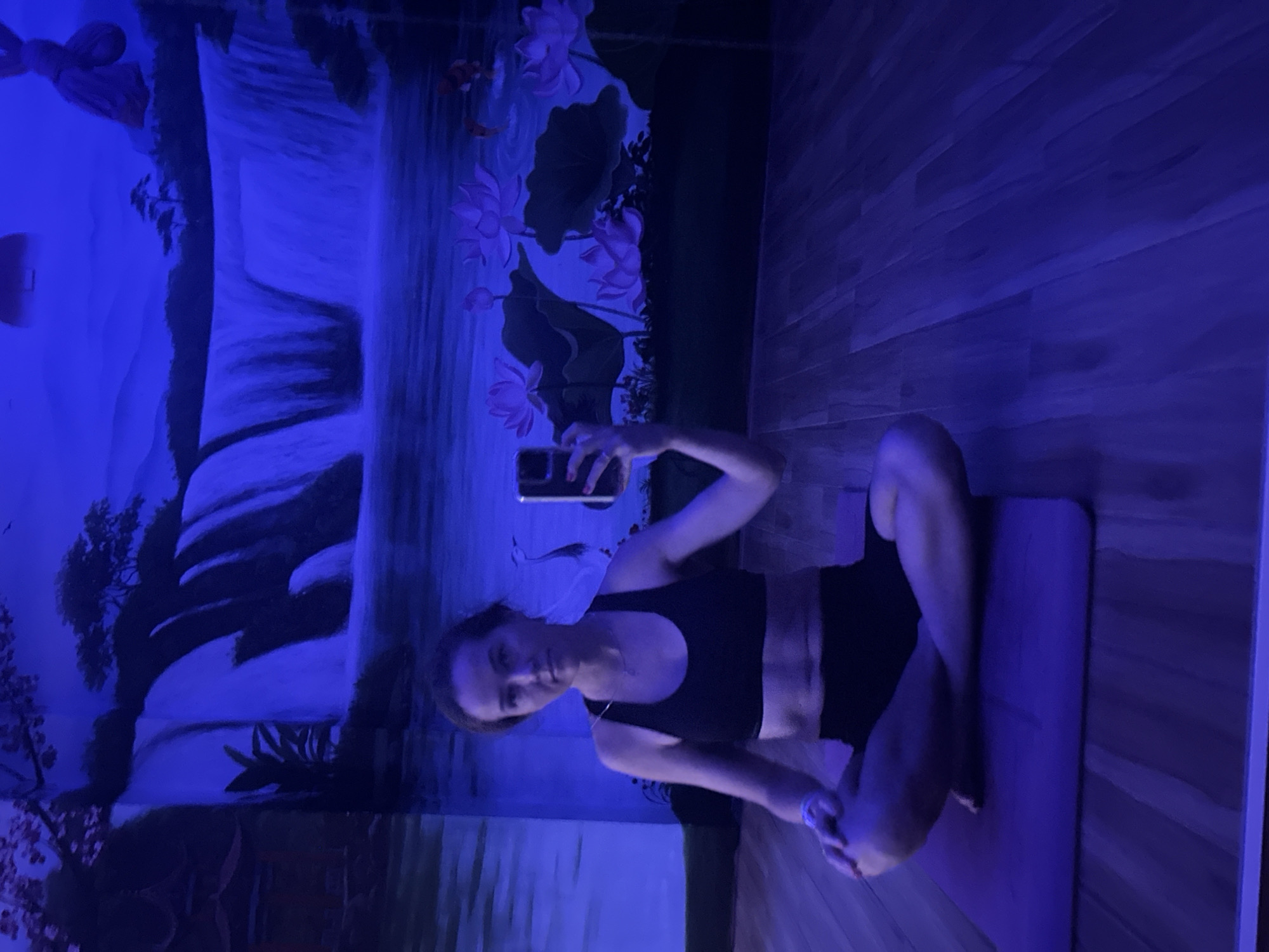 A woman sits cross-legged on a purple yoga mat in a room with a mural depicting a waterfall and lotus flowers. She wears a black sports bra and shorts and holds a cell phone in her right hand, presumably taking a photo or video. The room has wooden-look flooring. The lighting is primarily blue. This image was taken in Bac Nha Trang Ward, Vietnam.