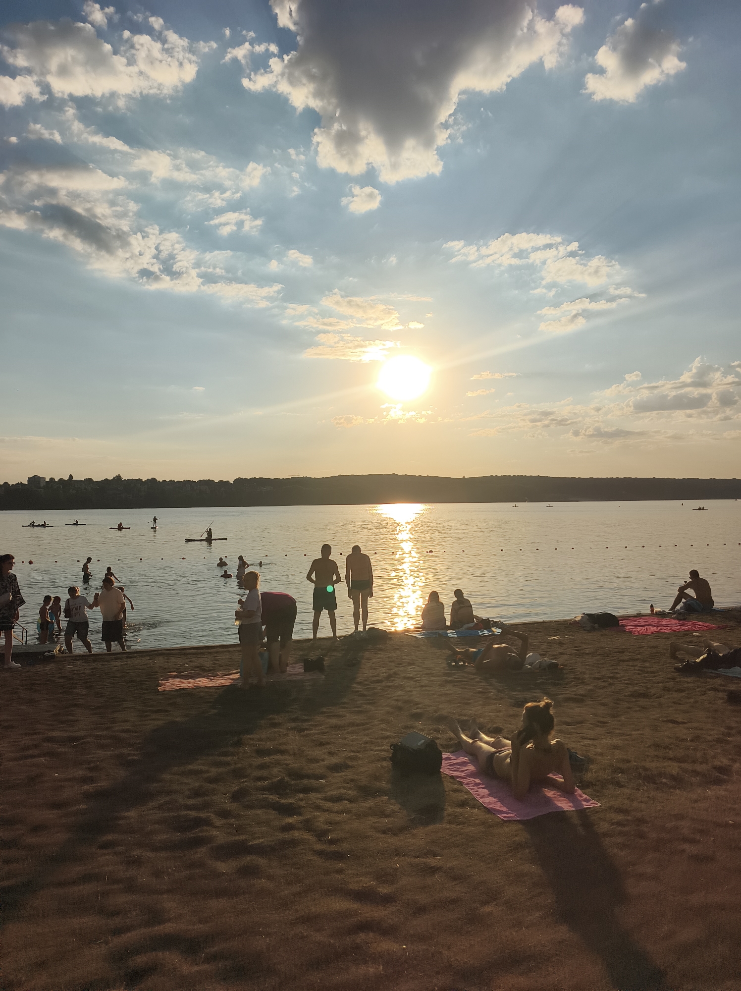 The image depicts a sunny day at a beach, likely Ternopil Lake in Ternopil, Ukraine, given the context. The sun is setting, casting a warm glow on the scene. People are engaged in various activities, including swimming, sunbathing, and paddleboarding. The beach is sandy, with some patches of grass. The water is calm and reflects the sky. In the distance, a treeline and buildings are visible on the horizon. The overall atmosphere is relaxed and leisurely.