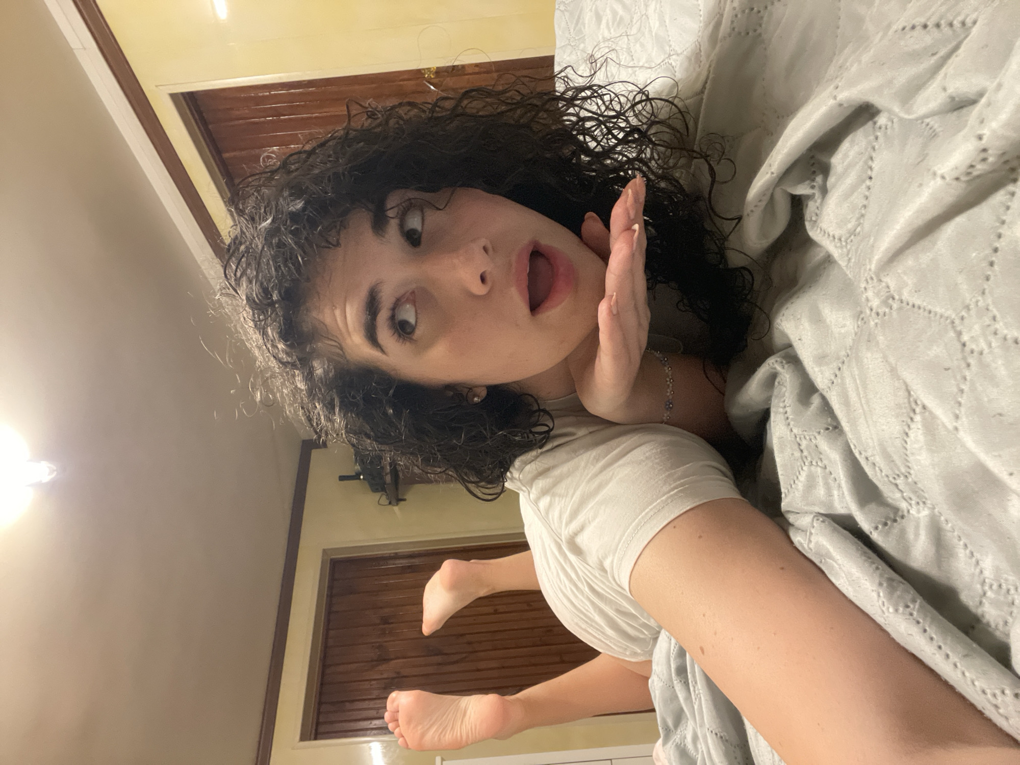 A young woman with dark curly hair is lying on a bed with a light gray textured comforter. She is propped up on her elbows, wearing a white t-shirt. Her face is turned to the right, with her mouth open in surprise or excitement as she looks upwards. Her right hand is held up to her chin. Her bare feet are visible, with the soles facing the camera. In the background, there are wooden doors and a yellow-painted wall, characteristic of a residential interior. The lighting suggests an indoor setting, possibly a bedroom. The scene does not provide any specific information that would indicate a location in Sofia, Bulgaria.
