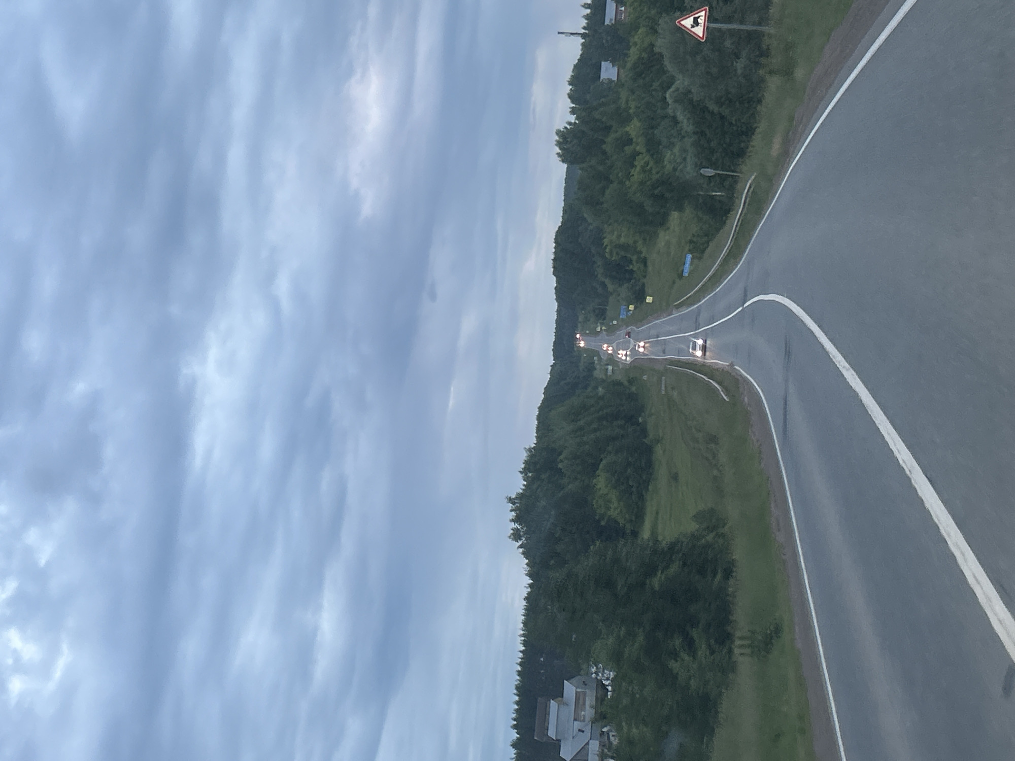 The image shows a two-lane asphalt road winding through a rural landscape under a cloudy, overcast sky. The road has white dividing lines and is bordered by grassy shoulders and dense green trees. Several cars are visible on the road, some with their headlights on, suggesting it may be dusk or a dark day.
On the left side of the road, there are several houses with pitched roofs. On the right side, there are more trees and a signpost with a triangular warning sign depicting a silhouette of an animal, likely indicating a wildlife crossing area. A few smaller signs are also visible along the roadside further down the road. The terrain appears gently rolling. The overall impression is one of a remote, scenic drive in a temperate climate.
The image does not contain any discernible elements that specifically place it in the city of Забегалово, Russia, beyond what might be inferred from the general appearance of rural Russian roadsides.