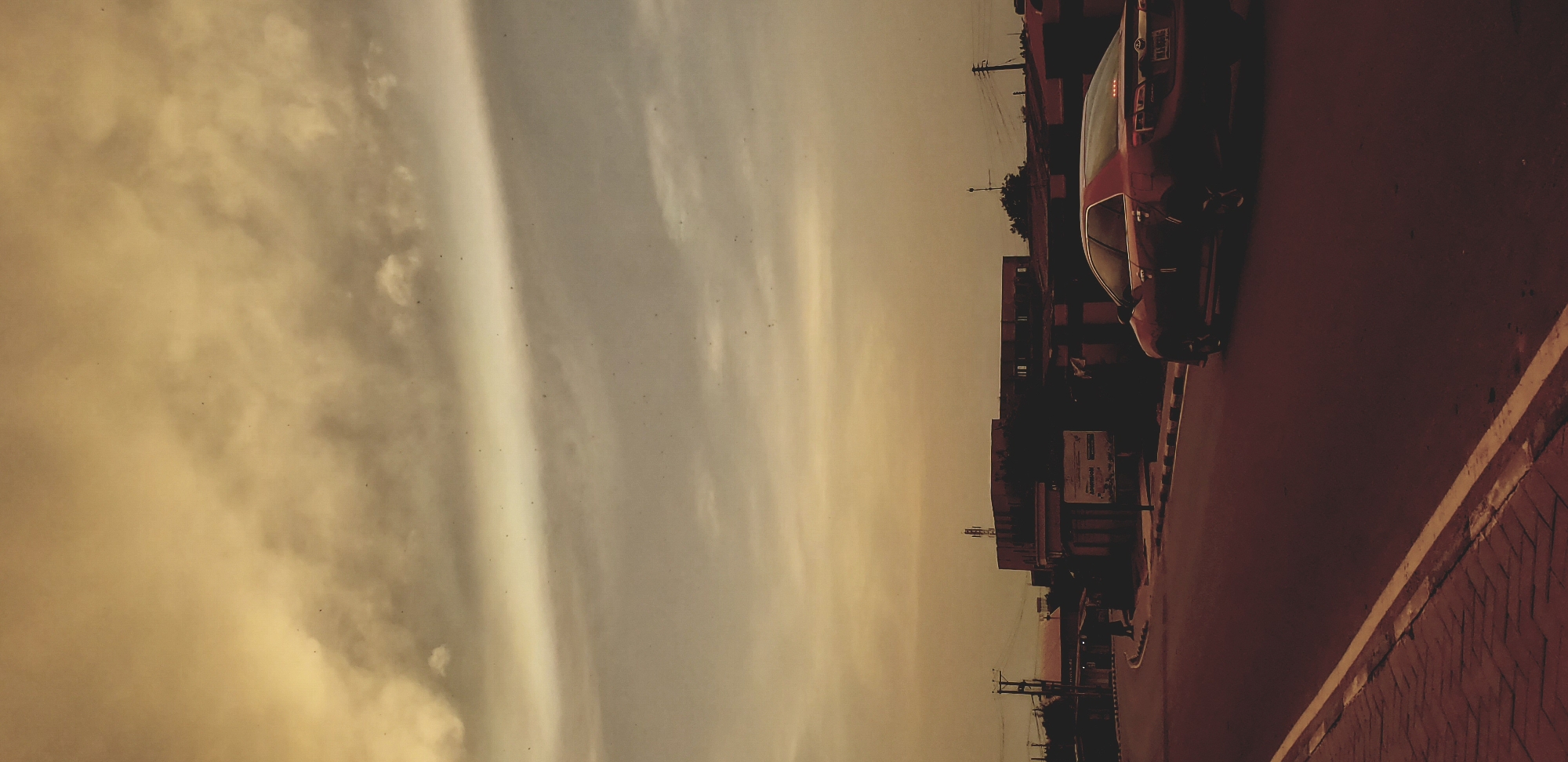 A street scene in Narayi, Nigeria, depicts a red car parked on the right side of the road. The sky is filled with clouds. Buildings and utility poles are visible along the roadside. The road surface appears to be paved.