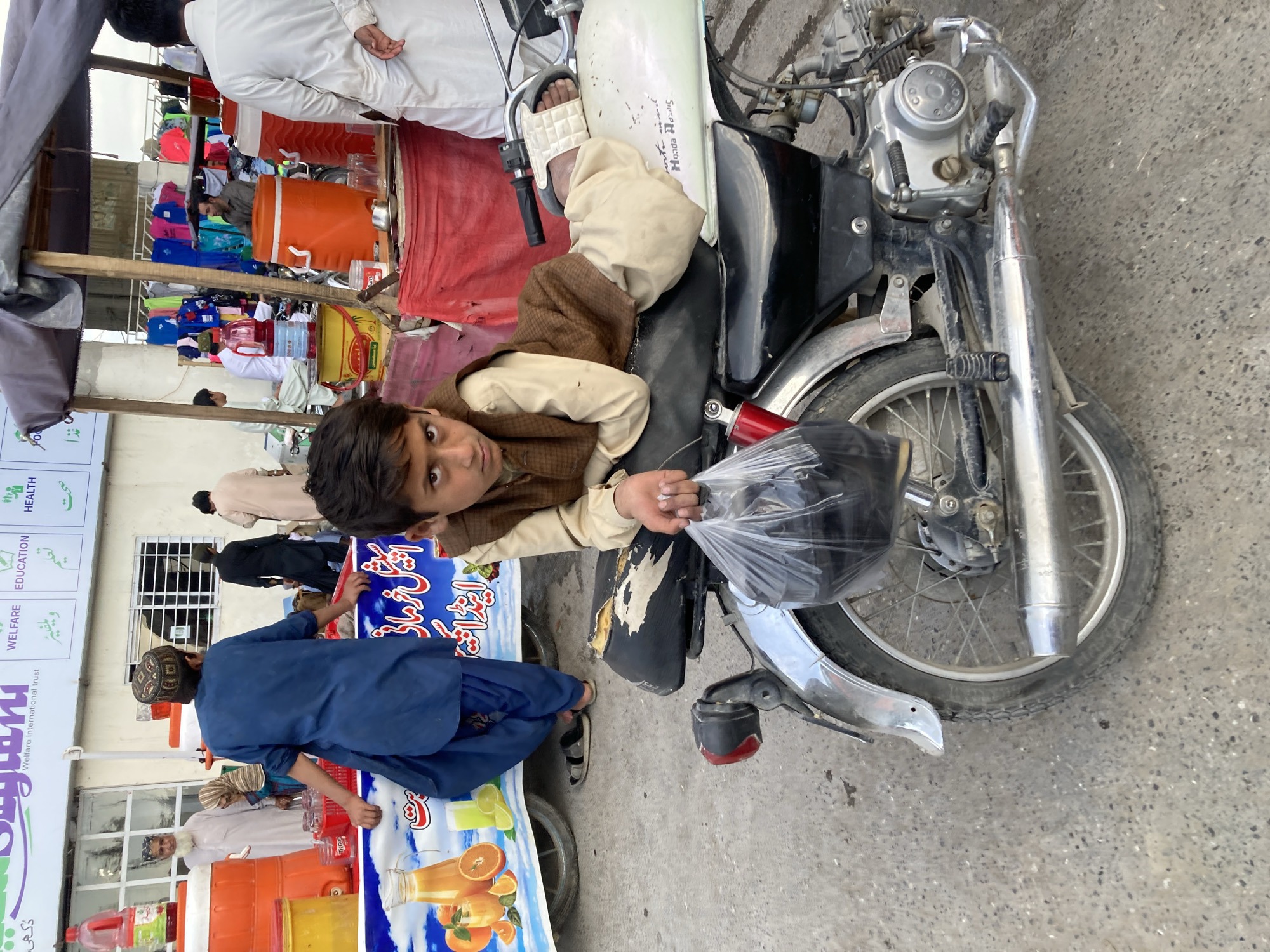 A boy in brown clothing lies on a motorcycle parked near a street vending stall in Quetta City Tehsil, Pakistan. He is holding a plastic bag. People are near the vending stall, and buildings and signage are visible in the background.