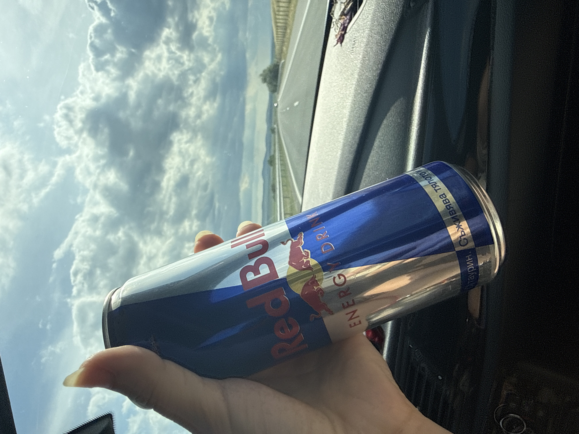 A hand holds a Red Bull energy drink can inside a vehicle. The can is blue and silver with the Red Bull logo visible. The background shows a highway stretching into the distance under a cloudy sky. The interior of the vehicle, including the dashboard, is visible in the foreground. The location is identified as Polsko Padarevo, Bulgaria.