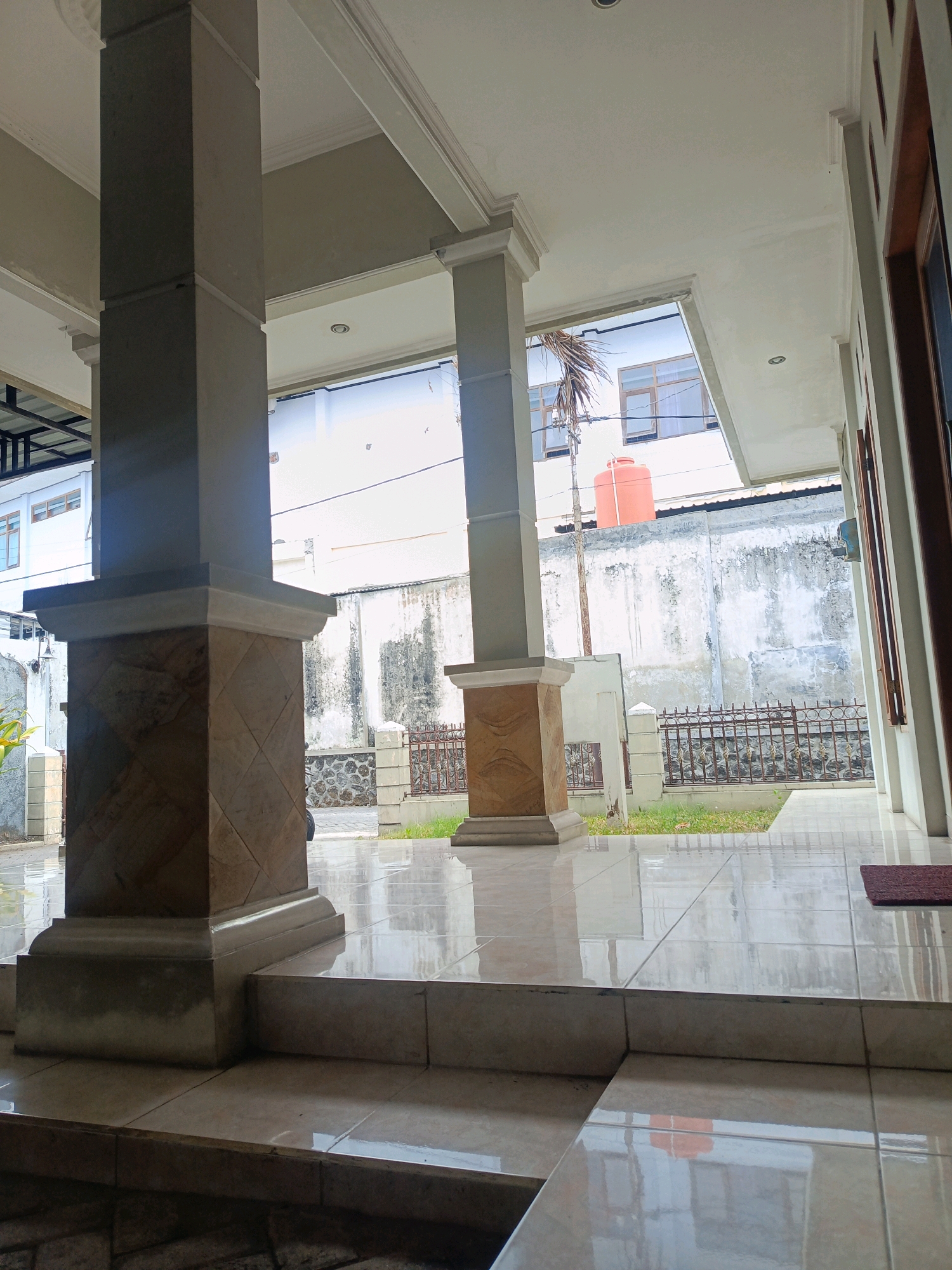 The image shows a covered walkway or porch with large square pillars, one of which is decorated with patterned tiles. The floor is tiled with large, glossy, light-colored tiles that reflect the environment. In the background, there is a wall with some discoloration and a rustic metal fence with decorative elements. Beyond the fence, there is a patch of grass and a building with multiple windows, and an orange water tank. The scene appears to be outdoors, in front of a residential or commercial building, possibly in the city of Kebonagung, Indonesia, given the architectural style and the presence of the water tank. There are no people visible in the image.