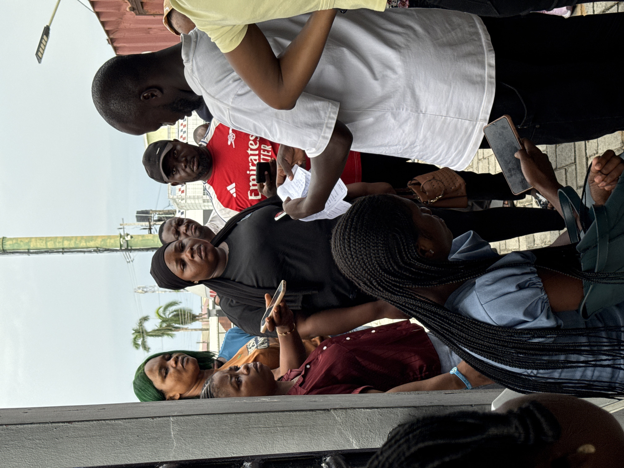 A group of approximately 10 people, mostly of African descent, are gathered outdoors in Ojomu, Nigeria. The setting appears to be an urban environment, indicated by buildings, palm trees, and overhead utility lines. A few individuals are looking at phones and papers, suggesting a potential transaction or consultation. Several people are standing close together. The image composition is a low-angle shot.