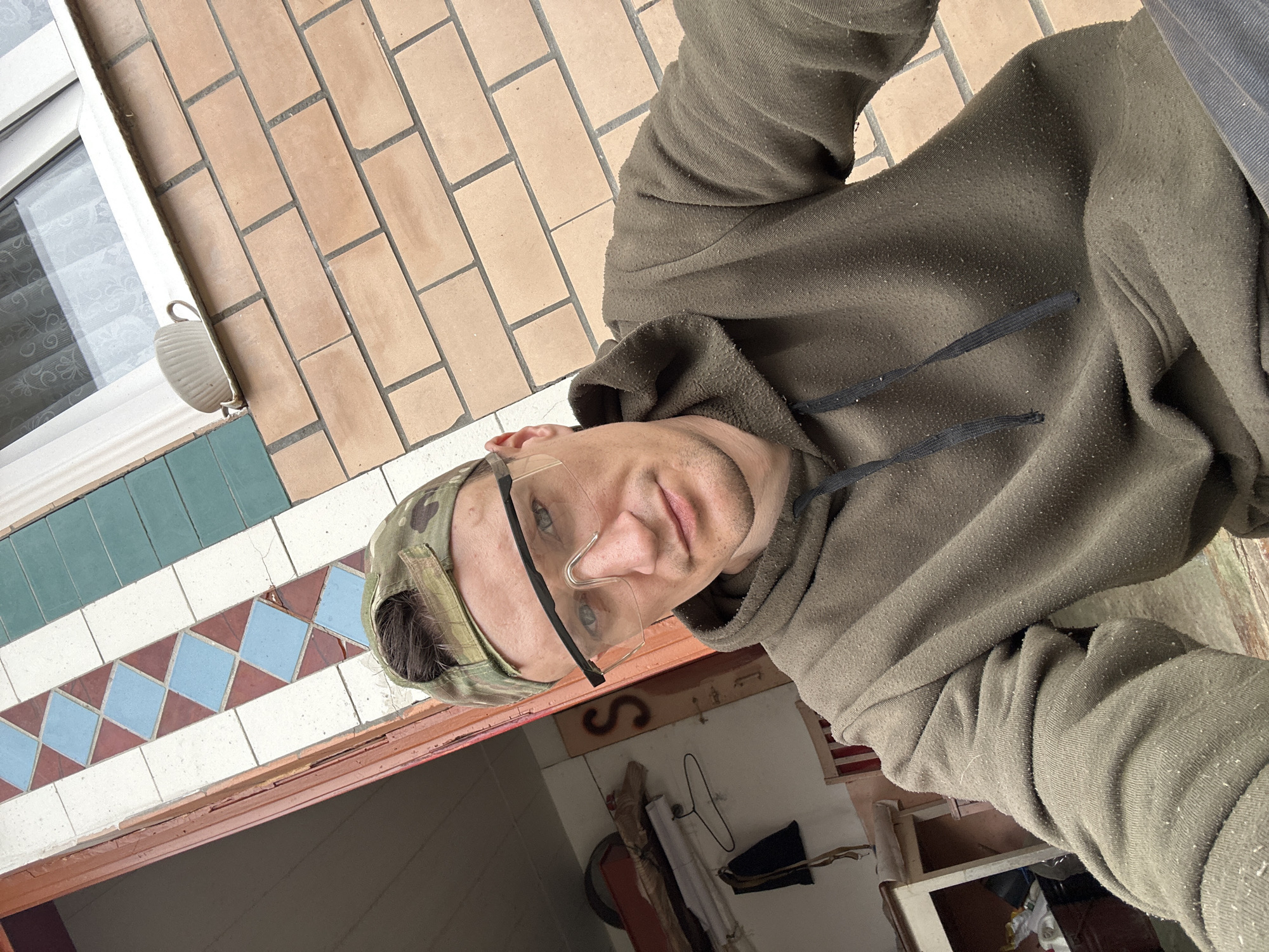 A man wearing a camouflage baseball cap and safety glasses is taking a selfie. He is wearing a dark green hooded sweatshirt. The background appears to be an outdoor setting, possibly in the city of Makiv, Ukraine. Behind him is a brick wall and a building with tiled decorative elements and a window. The man's face is visible, and he is looking towards the camera.