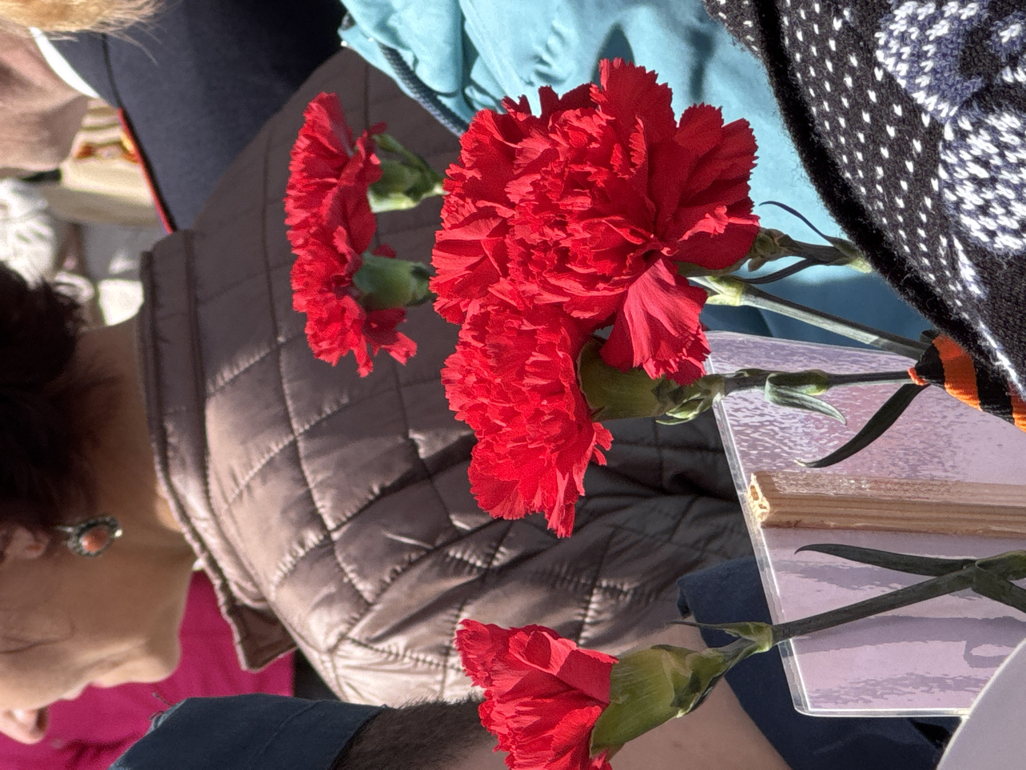 The image shows an arrangement of red carnations placed on a surface. A St. George ribbon is visible. Partial views of several people are present in the image, including a woman with an earring. The image was taken in Yamyshevo, Russia.
