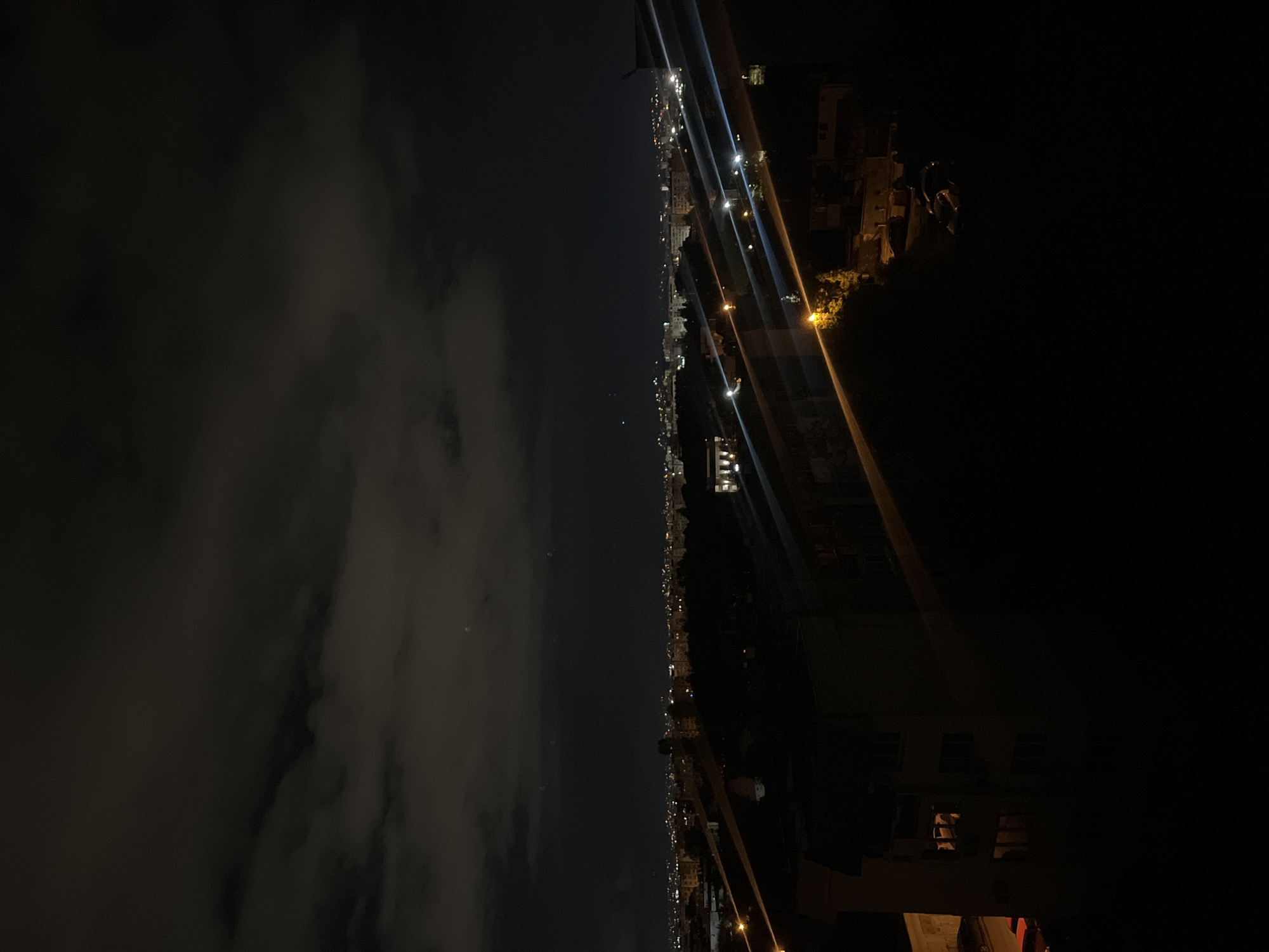 At night, the picture captures an elevated view of Sofia, Bulgaria. Dimly lit clouds fill the left side of the frame. Streetlights and building lights trace the cityscape, with some lights reflecting upward. On the right, partially obscured buildings suggest proximity to the vantage point. A light-colored building is partially visible in the foreground.