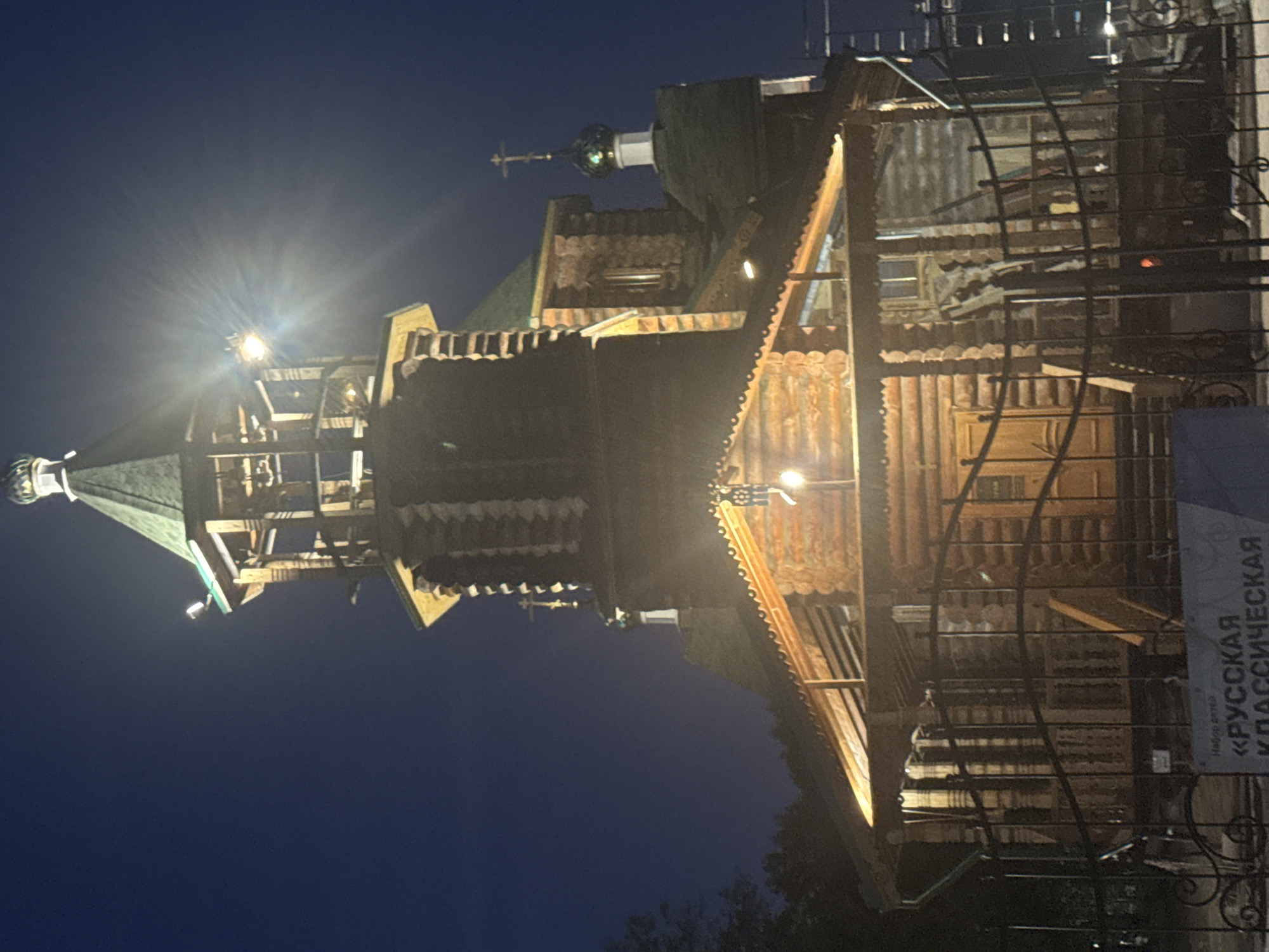 This image depicts a wooden church at night, illuminated by artificial lights. The church features multiple levels, a green roof, and a dome topped with a cross. The architecture suggests a traditional Russian style. A fence is visible in the foreground, and the background is a dark night sky with some trees on the left side. A sign with Russian text, possibly an advertisement or informational display, is partially visible at the bottom. The overall scene conveys a sense of tranquility and historical architecture set against the night.
