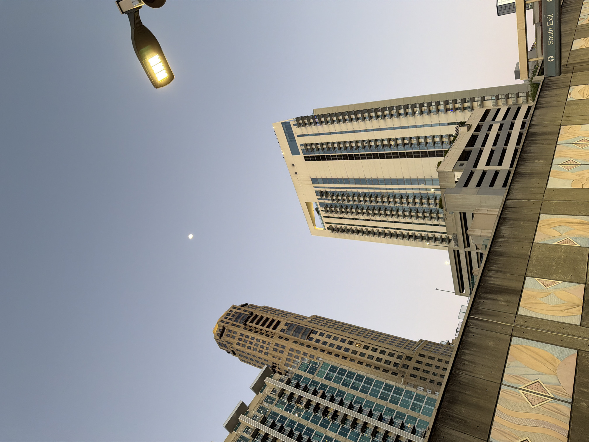 The image shows a view looking up at several tall buildings and a street light against a clear, pale blue sky. The buildings are modern and appear to be located in a downtown area, with one building featuring a grid-like pattern of windows and another with a more blocky, angular design. A street light is visible in the upper left corner of the frame, casting a warm yellow glow. The angle of the shot emphasizes the height of the structures and the vastness of the sky. The location appears to be near the CNN Center and the Georgia World Congress Center in downtown Atlanta, United States of America.