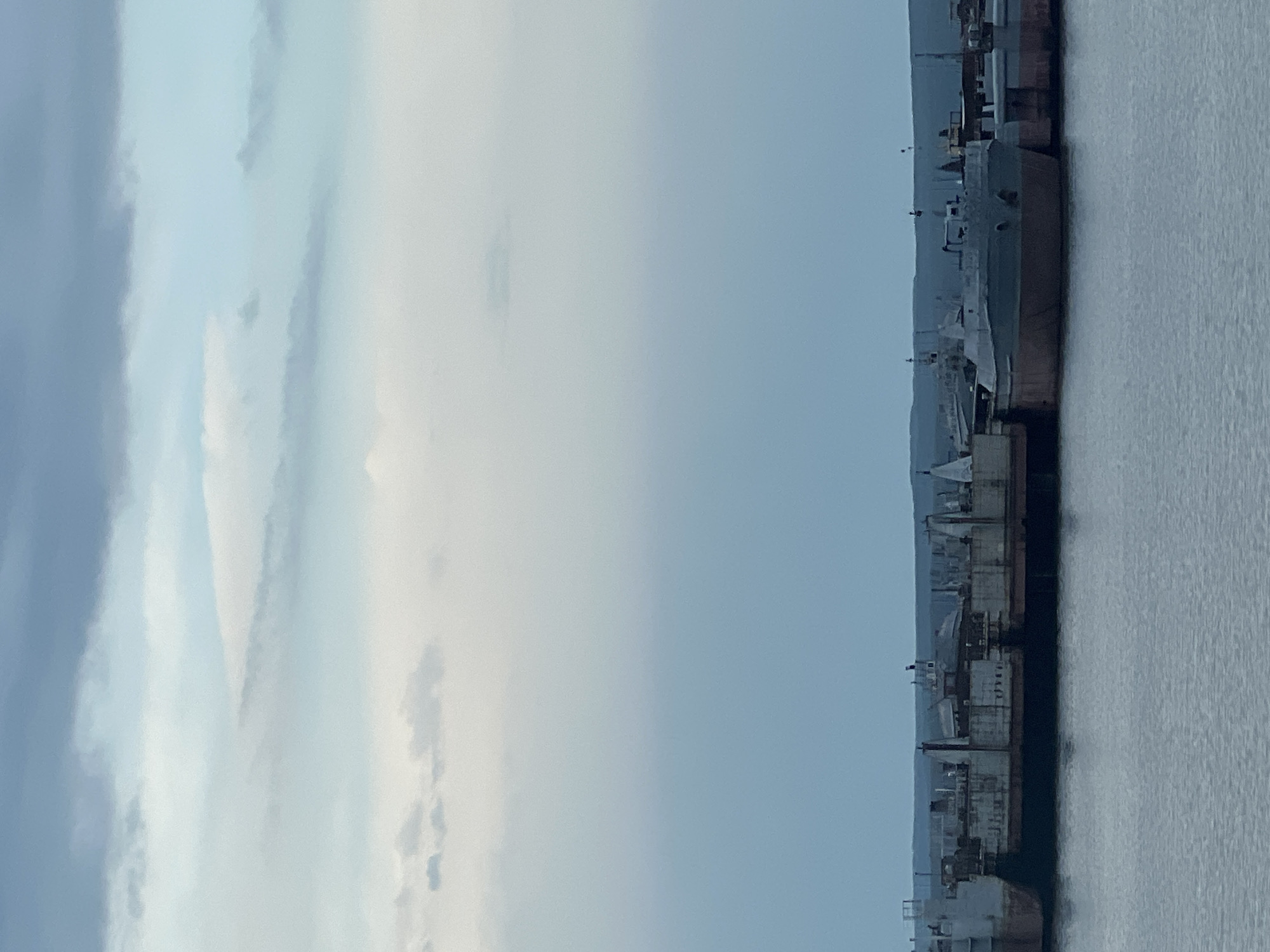 The image shows a view of a harbor in Chaikovsky, Russia. The right side of the image depicts a body of water, likely the Kama River. Along the water's edge are several docked ships or barges, varying in size and structure. Industrial equipment such as cranes and scaffolding can be seen along the harbor front. The sky takes up the left side of the image, showing a mix of light and dark clouds.