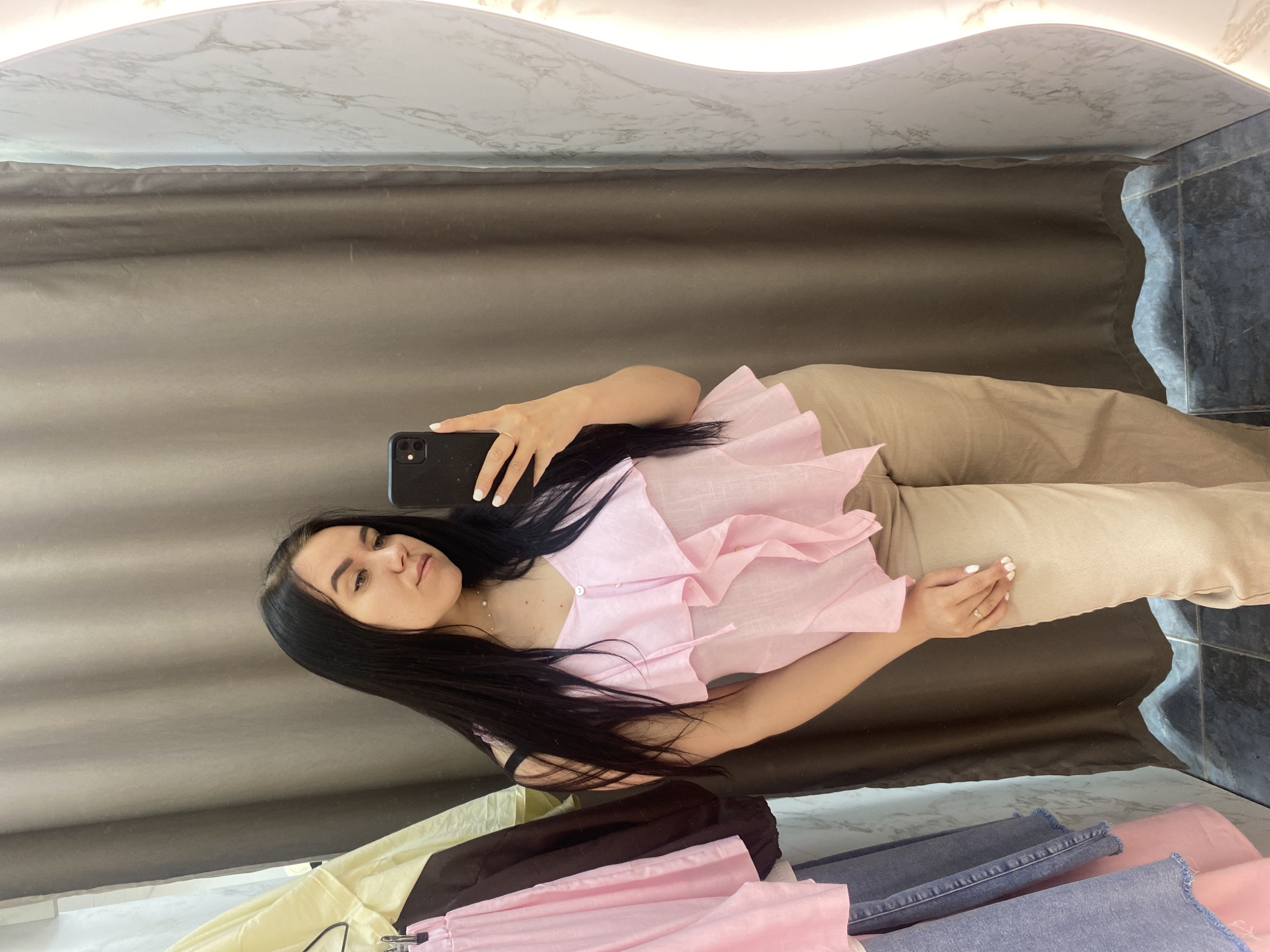 The image shows a young woman taking a selfie in a fitting room. She is holding a black smartphone in her left hand, wearing a pink ruffled top and beige pants. Her hair is long and black. Clothes are seen on the floor near her. The fitting room has a grey curtain as a backdrop and a marble-like surface on the top. The location is inferred to be in Chaikovsky, Russia based on user prompt.