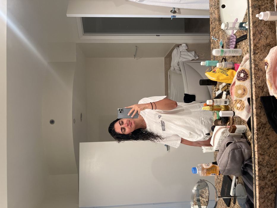 The image shows a woman standing in front of a bathroom mirror in a hotel room. She is wearing a white t-shirt and has her hair pulled back from her face. The woman is holding a smartphone and is looking at herself in the mirror. There are various toiletries on the counter behind the woman, including bottles of shampoo, conditioner, and lotion. There is also a washcloth with a donut pattern on it. The bathroom is tiled and has a white bathtub. The location is a hotel room in Miami Beach, Florida, United States. The coordinates, latitude 25.773 and longitude -80.141, place the scene in the heart of Miami Beach, known for its luxurious hotels and beaches.