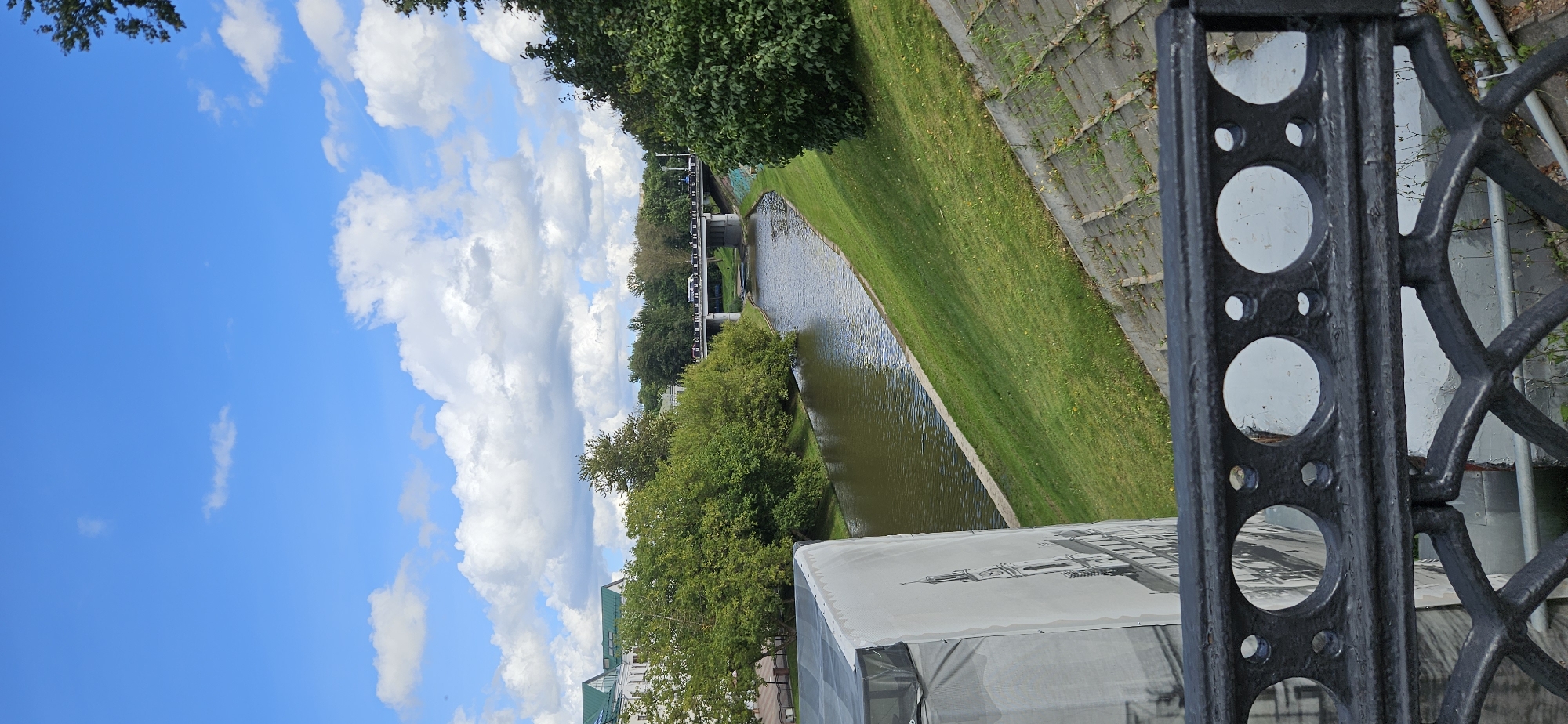 The image shows a view of the Western Dvina River in Vitebsk, Belarus, from a bridge or embankment. In the foreground, there is a decorative black metal railing with circular and oval openings. Behind the railing, a large white tarp is visible, partially covering a structure and displaying a black and white image of a building with a clock tower. The river flows through a grassy embankment to the right, with a concrete embankment on the far side. A bridge with vehicles crossing it spans the river in the middle ground. Lush green trees and buildings line the riverbanks. The sky is bright blue with fluffy white clouds. The scene appears to be a public space in a city park or along a waterfront promenade.