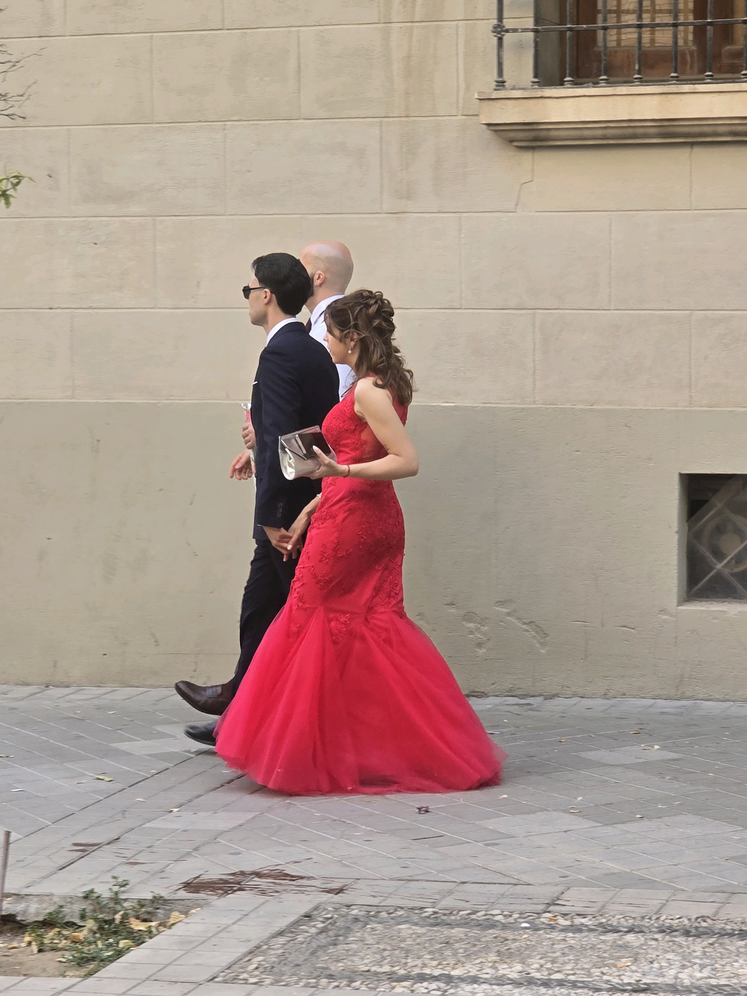 A group of three people are walking on a sidewalk in front of a beige brick building. The person on the left is a man wearing a dark suit and sunglasses, with his hands in his pockets. Next to him is another man, bald, wearing a white shirt and a dark tie. To the right of these two men is a woman in a bright red, floor-length gown. She is holding a silver clutch purse in her right hand and is walking with her left hand extended, possibly reaching for the man in the suit. The building behind them has a window with a dark metal grate. The sidewalk is made of light gray square tiles with a dark border. There are some dry leaves scattered on the ground. The image appears to be taken in Granada, Spain.