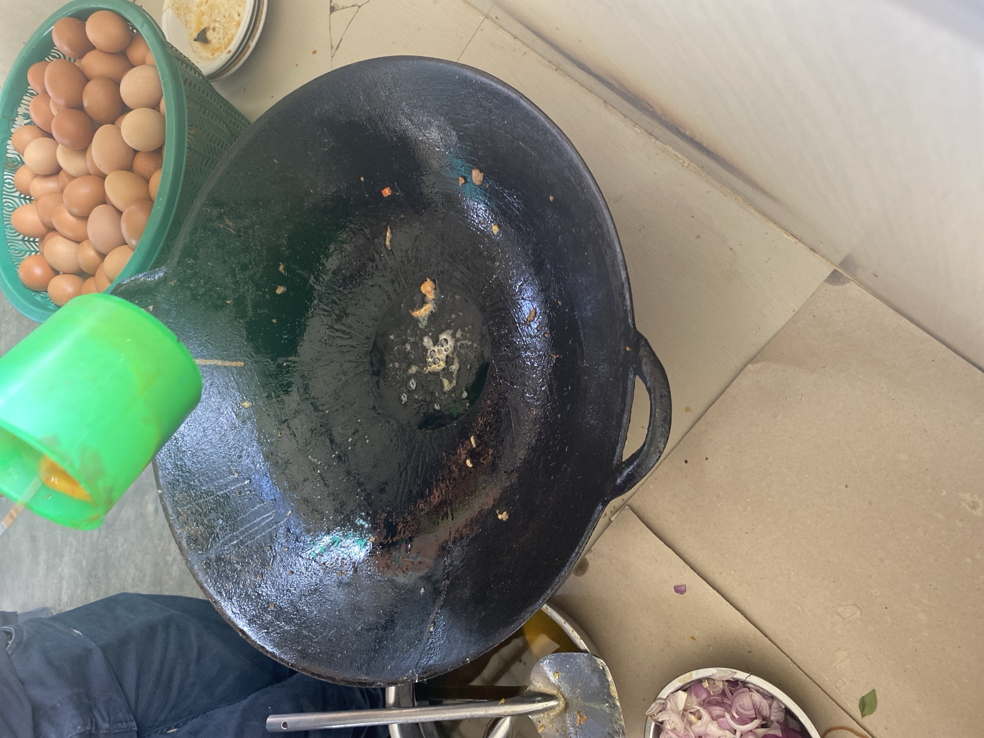 An overhead shot shows a large, seasoned black wok being prepared for cooking on a light-colored tiled surface. A person, wearing dark pants, is visible on the left, holding a green plastic cup from which a yellow liquid, likely beaten eggs, is being poured into the wok. Scattered around the wok are remnants of ingredients: a small pile of chopped red onions in a metal bowl on the lower left, and a basket filled with brown eggs is visible on the upper right. A metal spatula rests on the edge of a bowl containing some liquid and what appears to be fried food bits on the left side of the wok. The overall scene suggests food preparation, possibly in a street food stall or home kitchen. The location is identified as Banda Aceh, Indonesia.