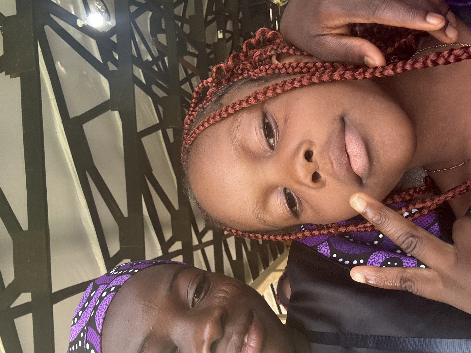 Two individuals, likely children, are captured in a close-up selfie. The person on the left, wearing a purple patterned headscarf and black clothing, is looking towards the camera with a slight smile. Their face shows some scarring on the left cheek. The person on the right, whose face is also partially visible, has their arm around the other individual. This person has braided hair with a reddish-brown hue. The background suggests an outdoor or semi-outdoor setting, possibly a covered structure with exposed wooden beams and a lightbulb visible overhead, consistent with a marketplace or gathering area in Itoga, Nigeria. The image is taken from a low angle.