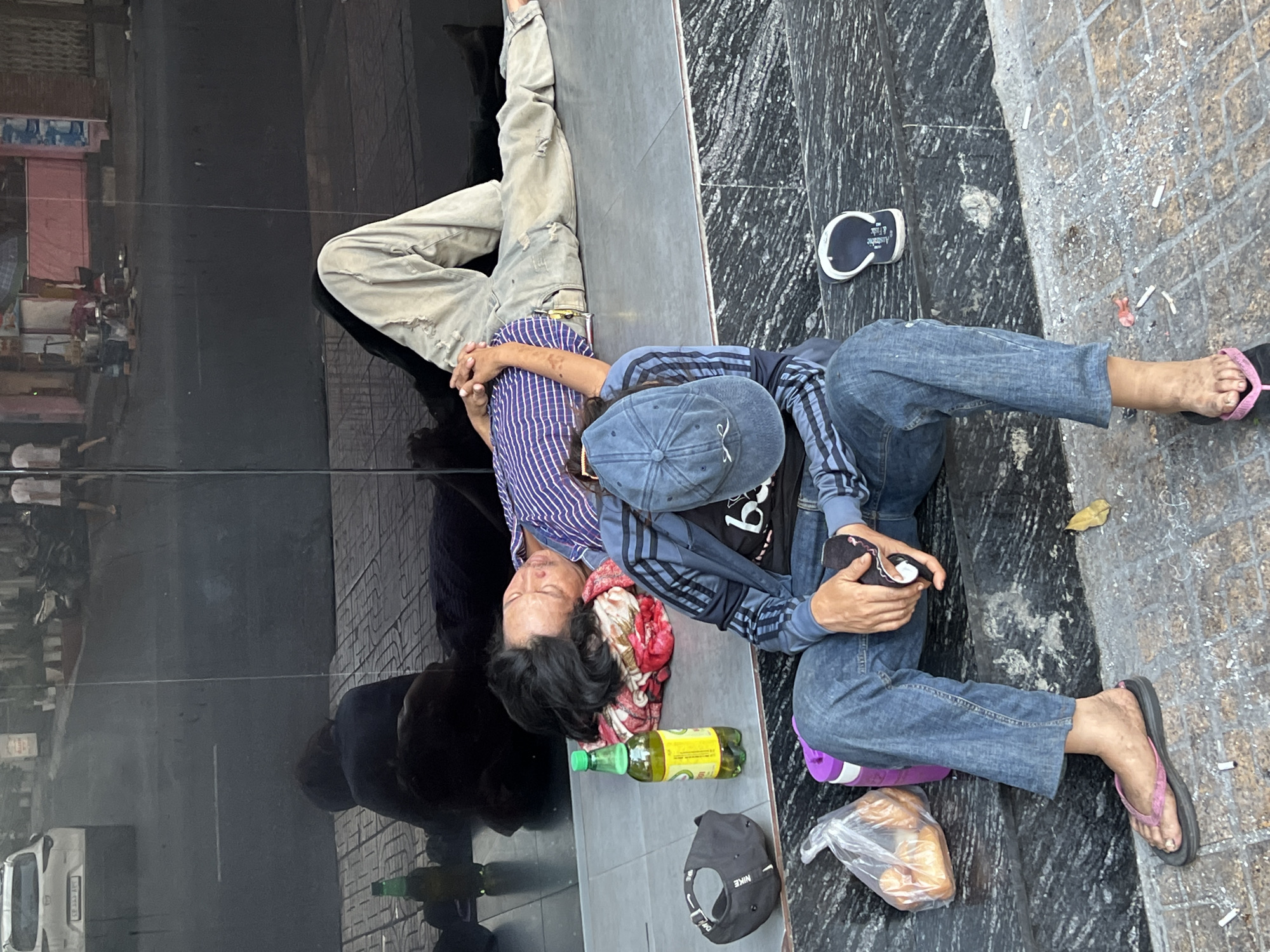 In Nha Trang, Vietnam, two individuals are observed sleeping on a tiled surface in front of a dark, reflective building. The person on the left is lying on their back, head resting on a colorful cloth, wearing a blue and white striped shirt. Beside them is a green bottle of liquid. To the right, another individual is lying on their stomach, with their legs extended. They are wearing light-colored, distressed pants and a dark top. In the foreground, a third person sits on the ground, facing away from the camera. They are wearing a blue jacket with black stripes, blue jeans, and a blue baseball cap. This individual is holding a black object in their hands and has a plastic bag containing bread, a purple thermos, and a black baseball cap nearby. Their feet are bare, one wearing a pink flip-flop. The ground is littered with cigarette butts. The overall scene suggests a public space where individuals are seeking rest.