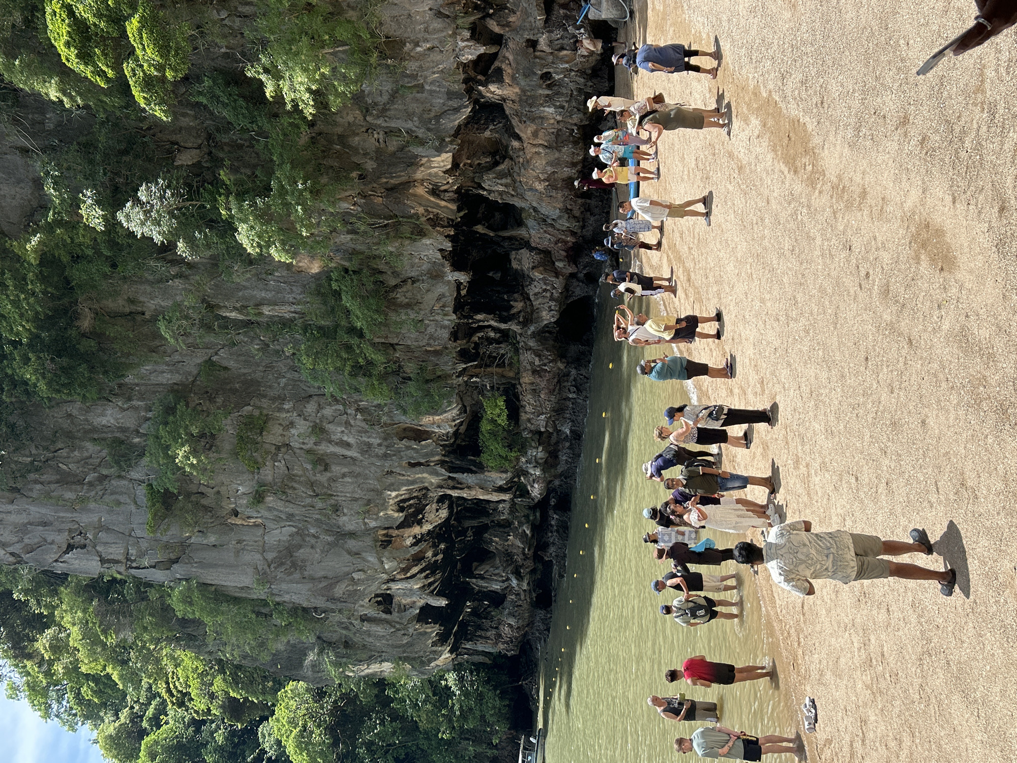 The image depicts a group of tourists on a sandy beach in Krasom, Thailand, with a towering limestone cliff face as a backdrop. The cliff is covered in lush green vegetation and features several large cave-like openings at its base. The water in front of the cliff is a calm, greenish hue, with yellow buoys marking a boundary.
The tourists, a diverse mix of ages and genders, are dressed in casual summer clothing. Some are standing in the shallow water, while others are gathered on the sand. Many are holding cameras or phones, seemingly taking pictures or videos of the scenery. A few individuals are engaged in conversation, while others appear to be listening to a guide or observing the surroundings.
In the distance, a small boat is visible near the shore. The overall atmosphere suggests a popular tourist destination, likely part of an island tour or excursion. The sunlight is bright, casting shadows on the sand and highlighting the textures of the cliff face and the foliage. The scene is tranquil and picturesque, characteristic of a tropical beach environment.