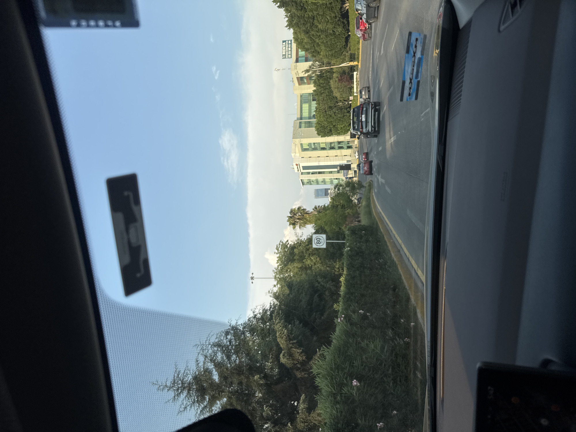 The image shows a view from inside a car, looking out onto a street in Puebla, Mexico. The road has multiple lanes and is bordered by trees and buildings. There are several cars on the road, moving in both directions. A sign displaying the number 80 is visible near the foliage. The buildings are multi-story structures, and the sky is partly cloudy. A sign with "RENTA" and a phone number is visible on one of the buildings. The image also contains elements of the car's interior, including the dashboard and windshield. A blue sticker is visible on the road with "LEXUS" written on it.