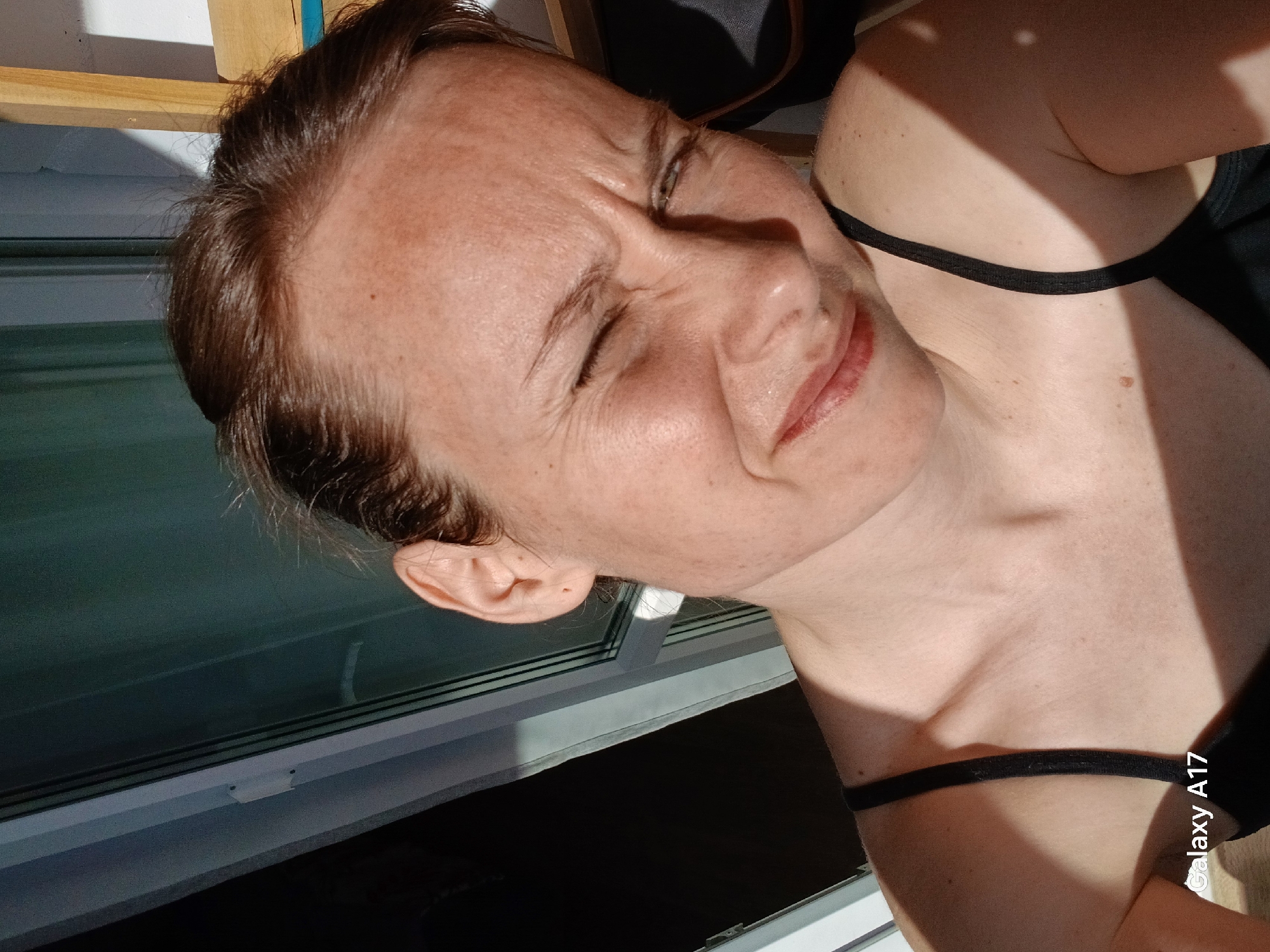 A woman is visible in a brightly lit indoor setting, likely near a window, given the strong light. She has fair skin, light brown hair, and is wearing a black spaghetti strap top. The lower portion of a window is visible in the background. "Galaxy A17" is visible at the lower right corner. The scene likely took place during daytime hours. The location cannot be verified as being in Yekaterinburg, Russia based on the image.