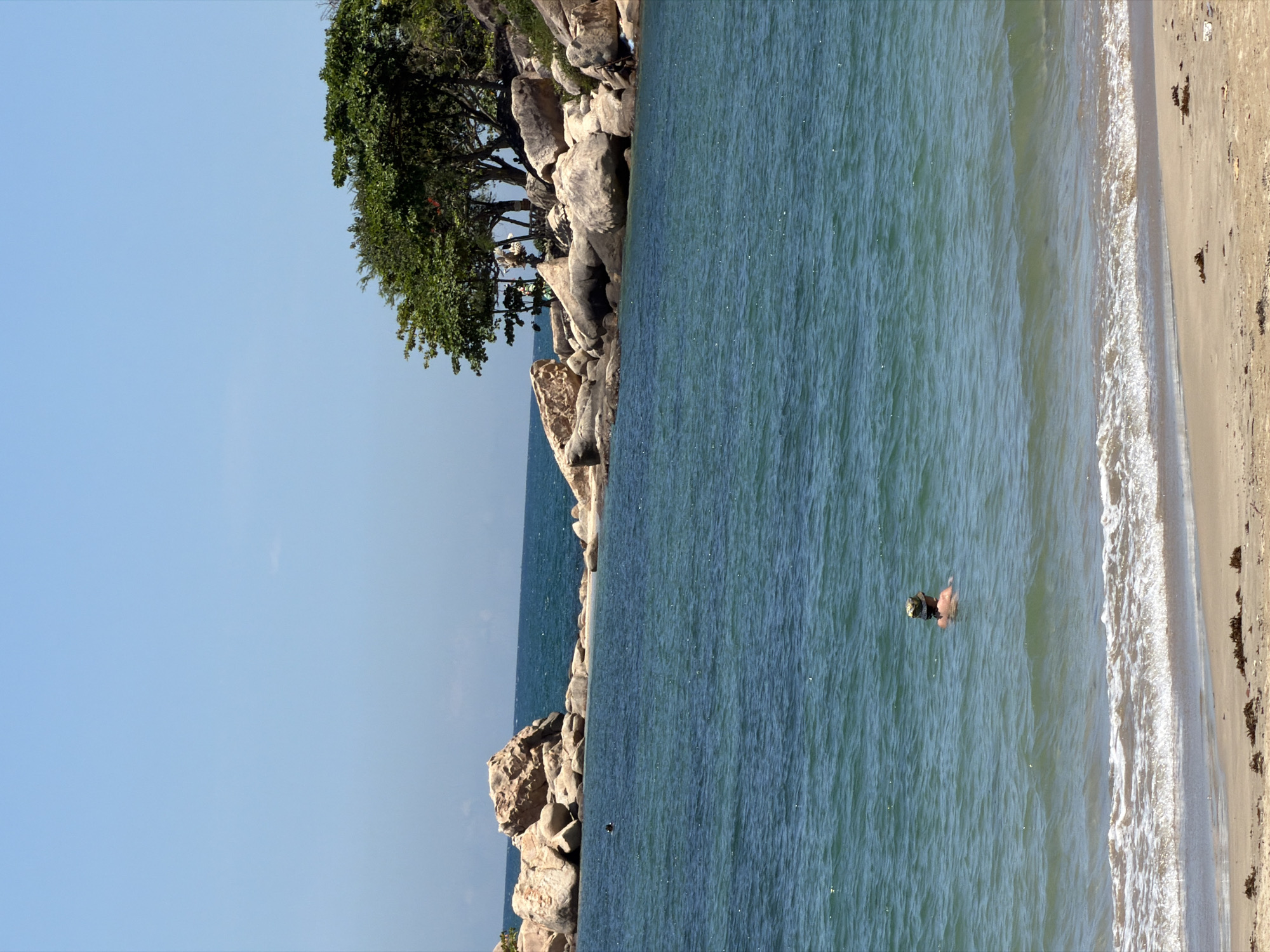 The image shows a coastal scene in Bac Nha Trang Ward, Vietnam. A rocky outcrop with trees lines the left side, transitioning into the sea. The water, with visible surface texture, meets a sandy beach on the right. A person is seen swimming in the water. The sky is a clear, light blue.