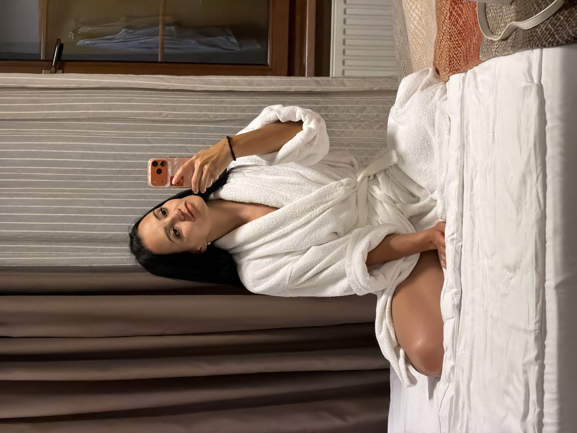 A woman is sitting on a bed in a room, taking a selfie with her phone. She is wearing a white bathrobe and has her legs crossed. The bed is covered in white linens. In the background, there are brown and grey curtains, a window, and a radiator. The overall impression is of a hotel room in Svetlogorsk, Russia.