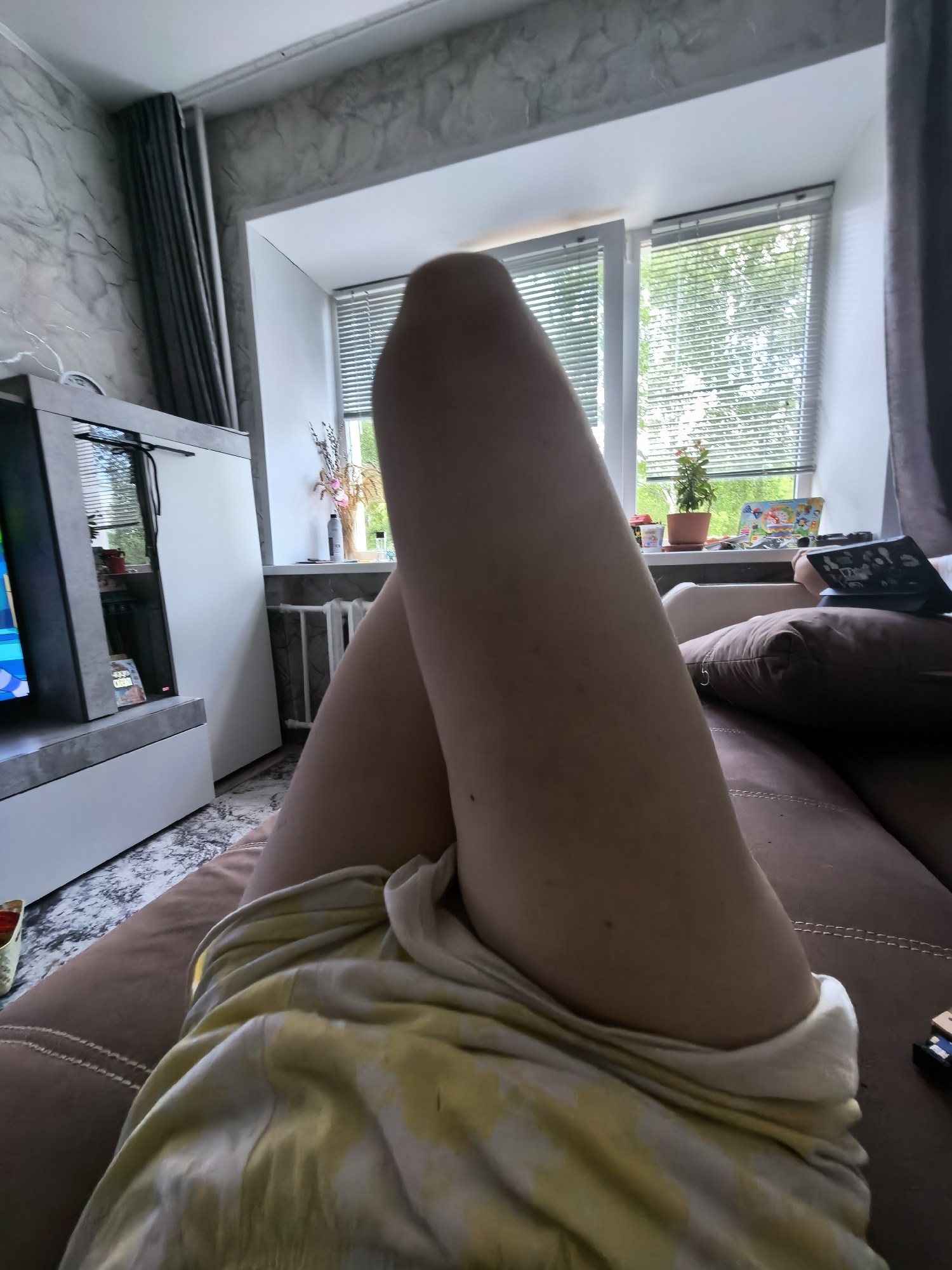 A first-person perspective image shows a person reclining on a brown couch indoors. The person's legs are crossed and angled towards the viewer, partially covered by a yellow and white patterned cloth. A television stand with a television is visible to the left, with a light-colored wall and curtains behind it. In the background, a window with closed blinds reveals a view of trees. The image appears to be taken in a residential setting; the specific location in Sterlitamak, Russia cannot be determined from the given image.