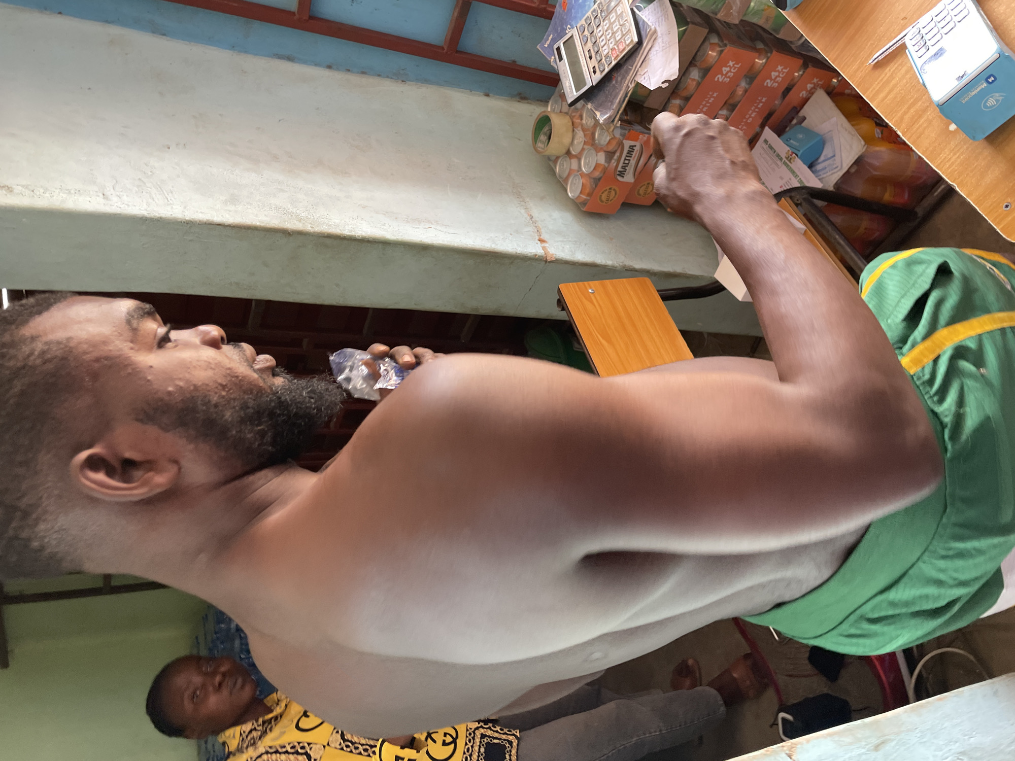 The image captures an indoor scene, likely a small shop or a home storage area, given the location context of Jos, Nigeria. In the foreground, a man with a beard is depicted shirtless, wearing green shorts or a similar garment. He is looking upwards and to the right, in the process of drinking from a clear plastic sachet or small bottle.
Above the main subject, on a concrete shelf, various items are stored. These include a calculator, several orange boxes of "Maltina" non-alcoholic malt drink, which are labeled "24 x 33CL" and state "NON-ALCOHOLIC MALT DRINK." A roll of packing tape and some papers are also visible on this shelf. To the right, on a light wooden table, a blue and white POS terminal with "Momo agent" clearly visible on its side is present, alongside a pen.
In the background to the left, another person, possibly a male, is partially visible, wearing a yellow patterned shirt and looking towards the camera. The lighting suggests it is daytime. The presence of Nigerian brands like Maltina and the Momo agent POS terminal are consistent with the provided location context.