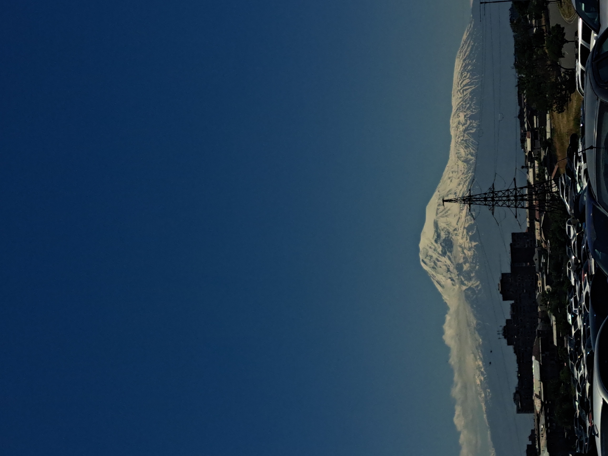 The image shows a view of Mount Ararat from Yerevan, Armenia. An electrical tower is visible in front of the mountain. A row of cars and buildings are also visible on the right side of the image. The sky is clear and blue.