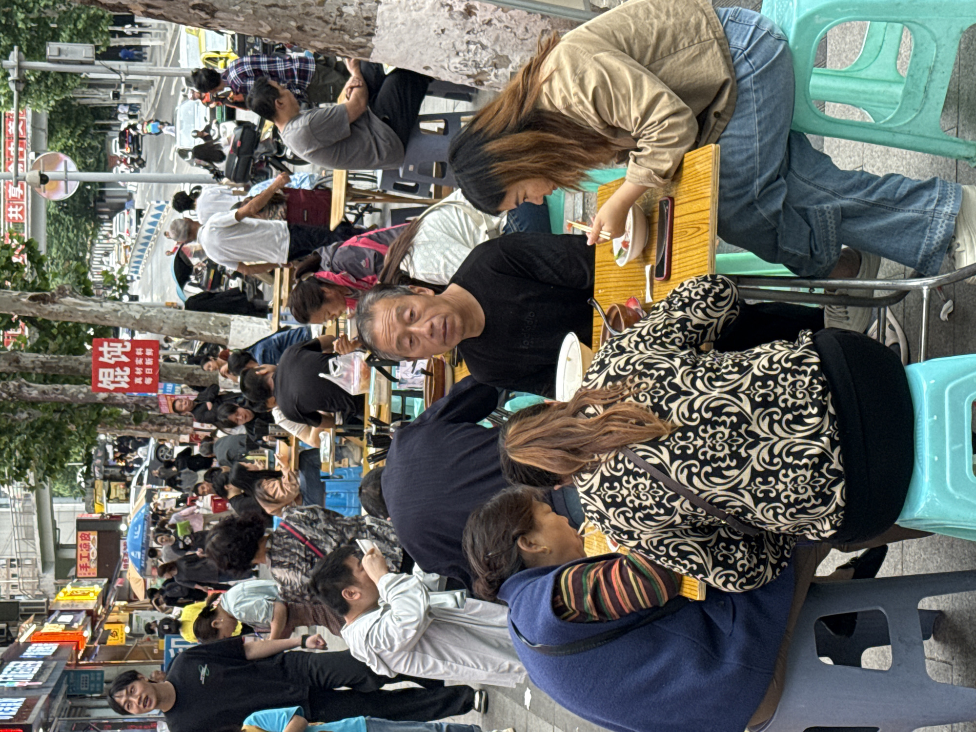 The image depicts a densely populated street scene in Yuzhong District, China. The foreground shows multiple people seated at low tables, seemingly eating or socializing, with a variety of dishes and items visible on the tables. Seating consists of simple plastic stools in light blue and dark gray. The individuals vary in age and attire, with some directly facing the camera. The background displays a bustling street extending into the distance, filled with pedestrians and vehicles. Storefront signs are visible along the street, with one sign in Chinese featuring red characters. The overall environment appears to be a busy commercial or dining area, with trees lining the street.