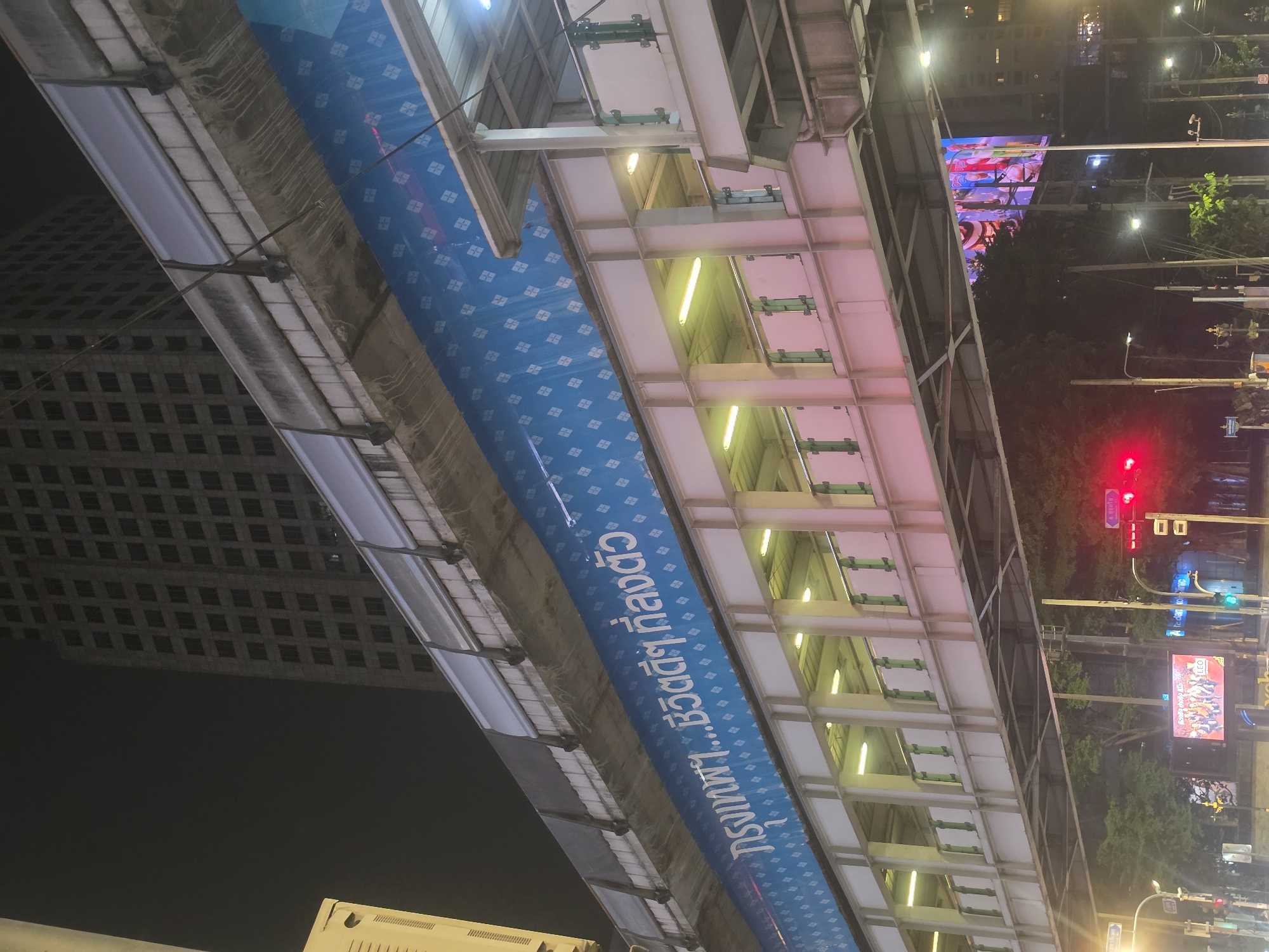 The image shows an elevated walkway in Bangkok, Thailand at night. The walkway is made of steel and concrete, with rows of rectangular openings. The walkway is illuminated with yellow lights. A blue banner with white text in Thai runs along the side of the walkway. In the background, there is a tall building with many windows. Streetlights, trees, and a billboard are also visible. Traffic lights are also visible.