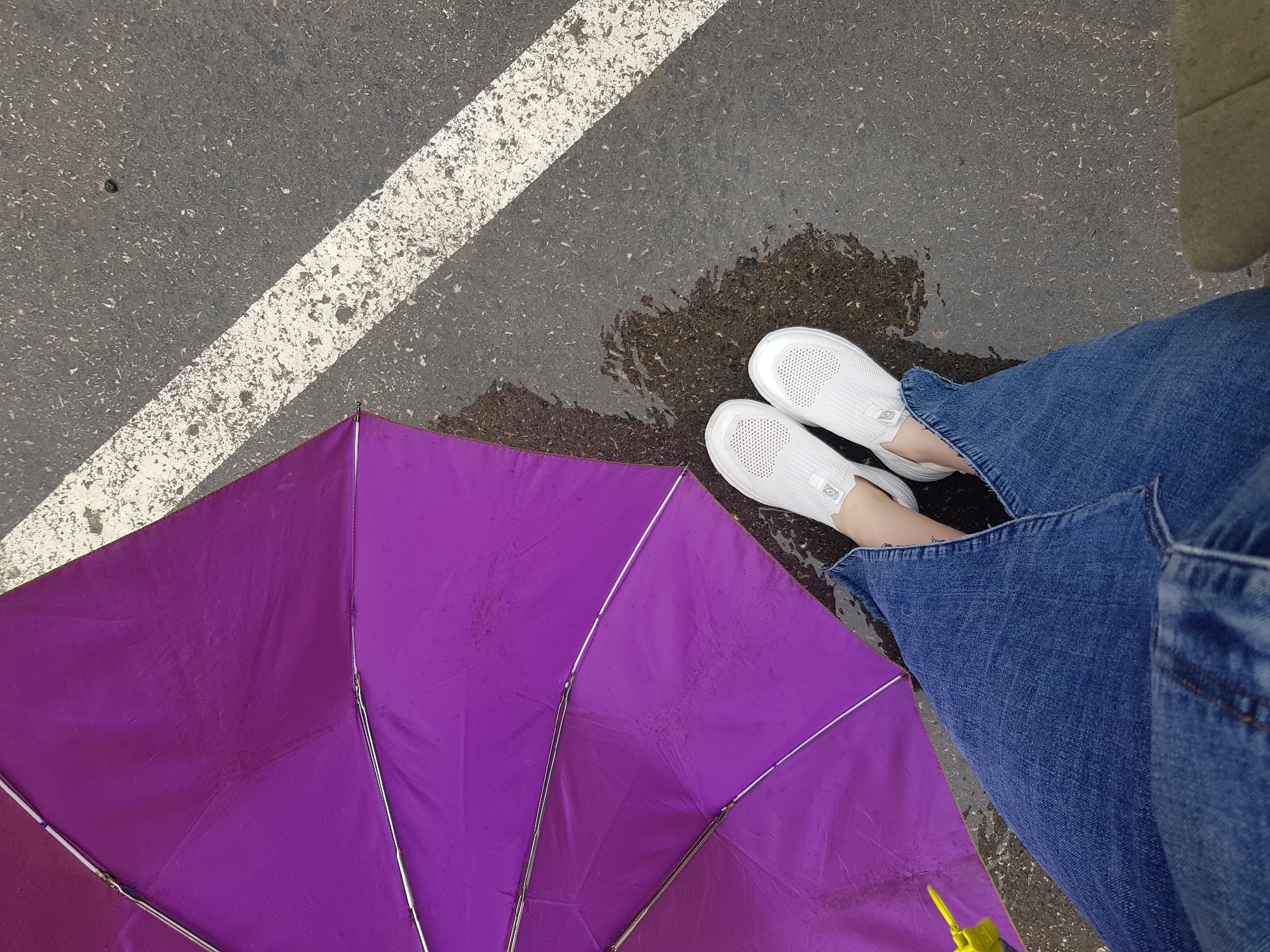 A first-person perspective shot captures a person standing on a wet asphalt surface, partially shielded by an open purple umbrella. The person is wearing blue denim jeans and white slip-on sneakers. The ground is speckled with light-colored debris and marked by a diagonal white line, typical of a road or parking lot. Puddles of water are visible on the asphalt, reflecting the surroundings. A small yellow object, possibly part of the umbrella or another item, is visible on the left side of the frame. The scene suggests a rainy day in an urban environment.