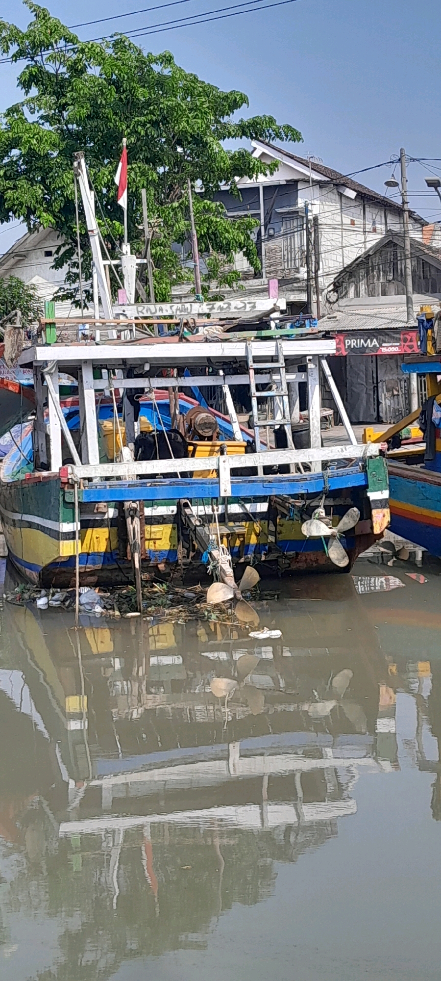 In Pasuruan, Indonesia, a weathered fishing boat is docked in a murky waterway. The boat, painted in vibrant stripes of yellow, blue, and green, shows signs of wear and tear. Debris, including plastic waste and vegetation, collects around its stern and in the water. A propeller is visible near the water's surface, partially submerged. Behind the boat, buildings with corrugated metal roofs line the bank, and lush green trees provide a natural backdrop. A flag of Indonesia flies from a mast on the boat. The water reflects the boat and the surrounding environment, creating a distorted mirror image.