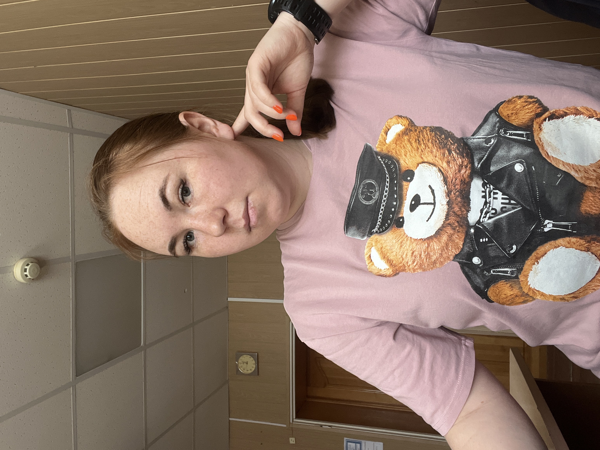 A woman is in an interior space with a tan tiled wall, brown striped ceiling, a clock, and a smoke detector. She wears a pink shirt with a bear in a black leather jacket on the front and a black watch on her wrist. Her nails are painted orange. The location is Chaikovsky, Russia.
