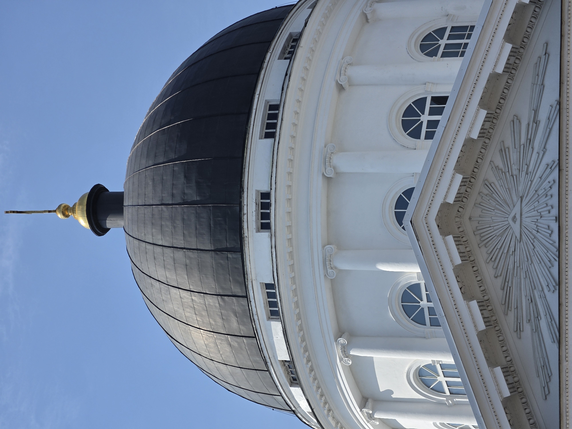 The image shows a close-up, angled view of the black dome of a white building against a clear blue sky. The dome has a metallic sheen and is segmented by dark lines. At the apex of the dome, a golden spire with a cross-like ornament is visible. The building's white facade features a series of classical architectural elements, including rounded columns with ornate capitals, decorative molding, and arched windows with segmented panes. Below the dome, there is a prominent pediment adorned with a sunburst motif and an all-seeing eye. The building is likely a religious or governmental structure. The image is a photograph taken from a slightly elevated perspective, looking upwards towards the dome. This building is the Kremlin's Archangel Cathedral in Nizhny Novgorod, Russia.